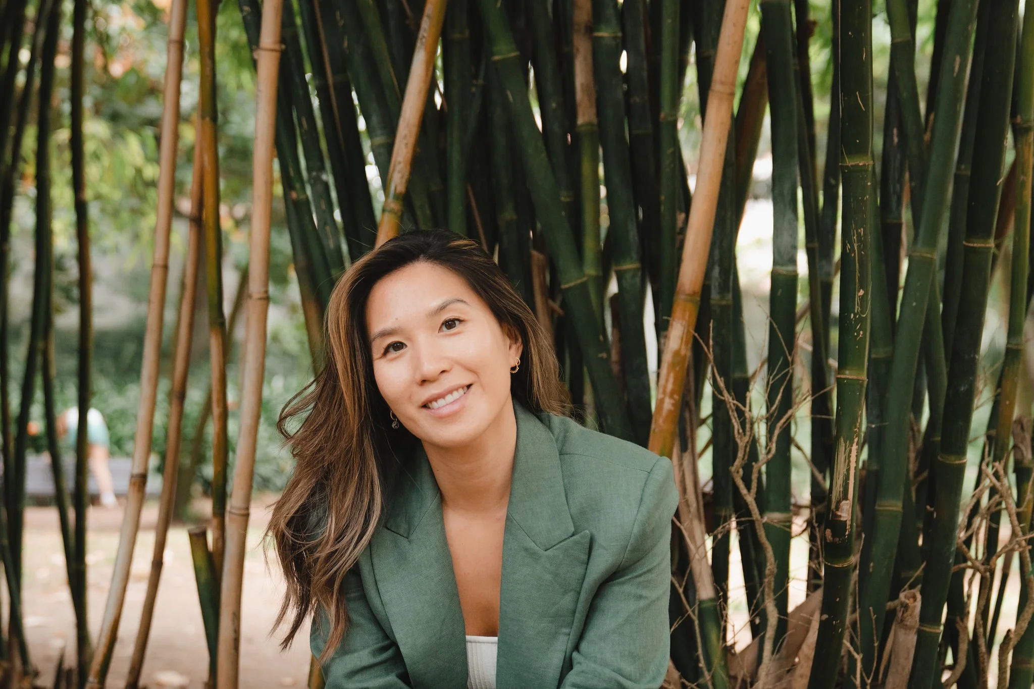 A woman with long wavy brown hair and a bright smile wearing a green blazer, sitting in front of tall bamboo stalks outdoors.