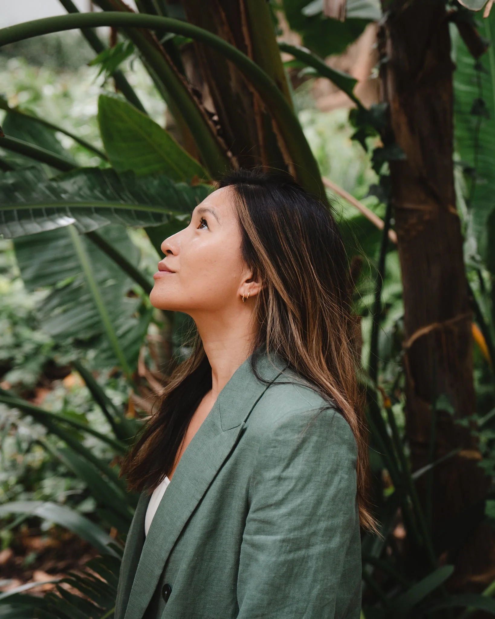 A woman with dark brown hair highlighted with blonde wearing a green blazer, looking upward in a lush jungle with large green leaves.