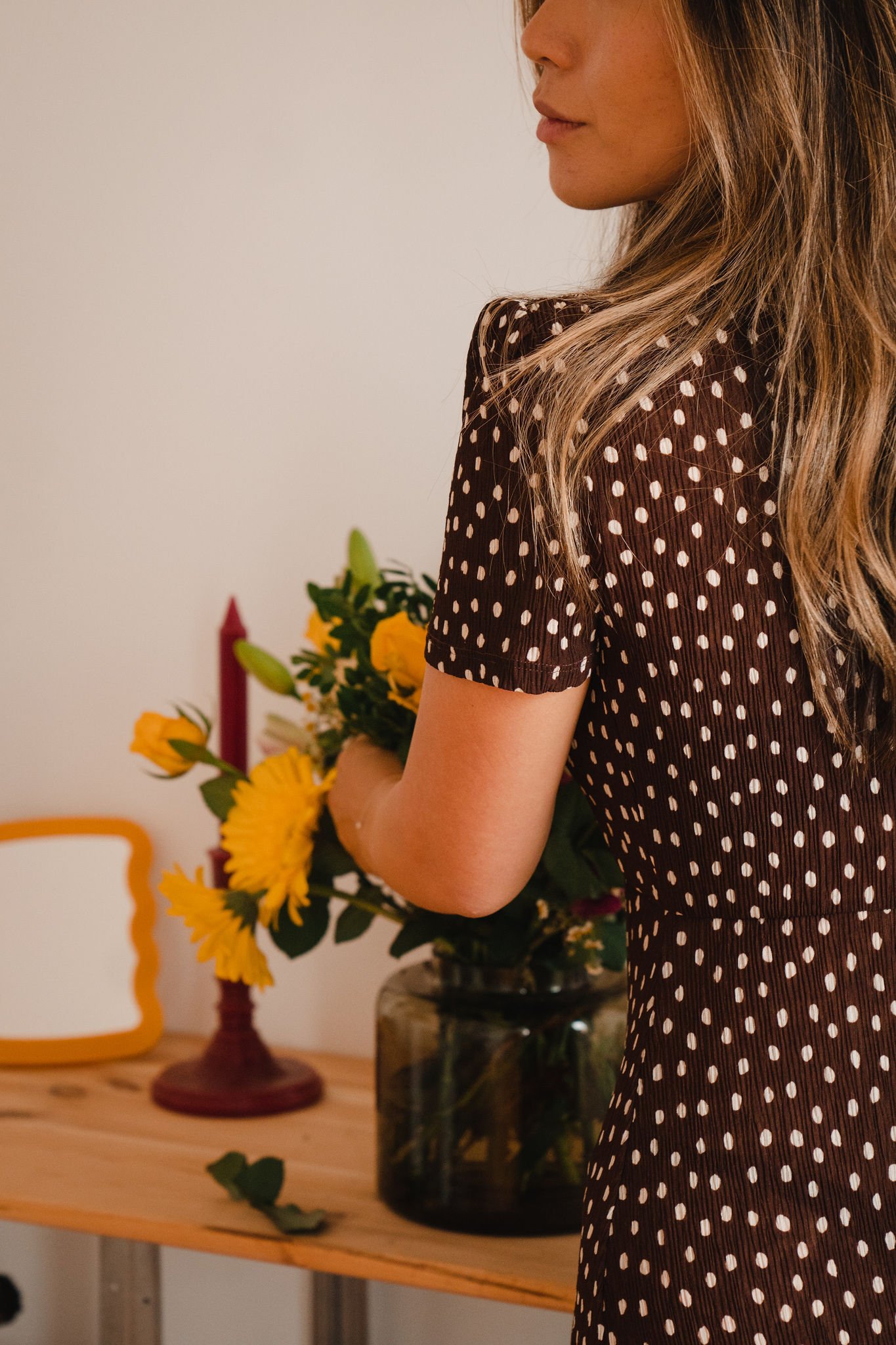 A woman with long, wavy blonde hair wearing a brown dress with white polka dots, holding a bouquet of yellow flowers.