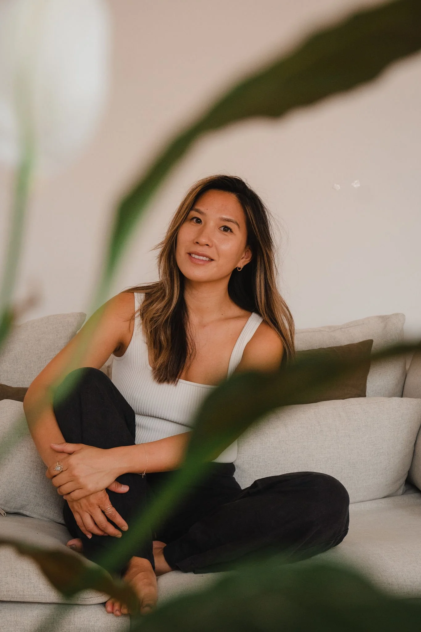 A woman sitting on a beige couch, smiling at the camera, wearing a white tank top and black pants, framed by green leaves in the foreground.