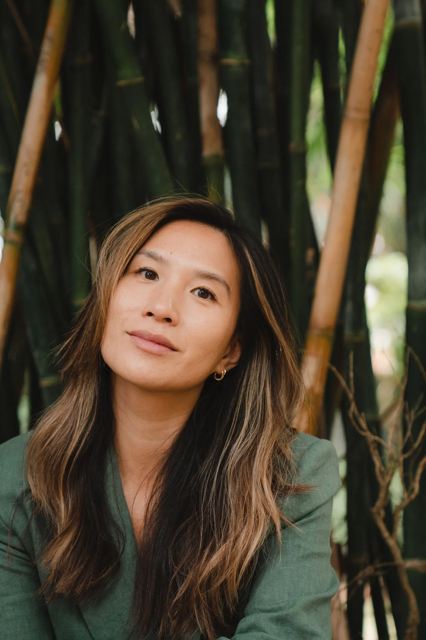 A woman with long, wavy hair and hoop earrings, wearing a green blazer, posing outdoors with tall green plants in the background.