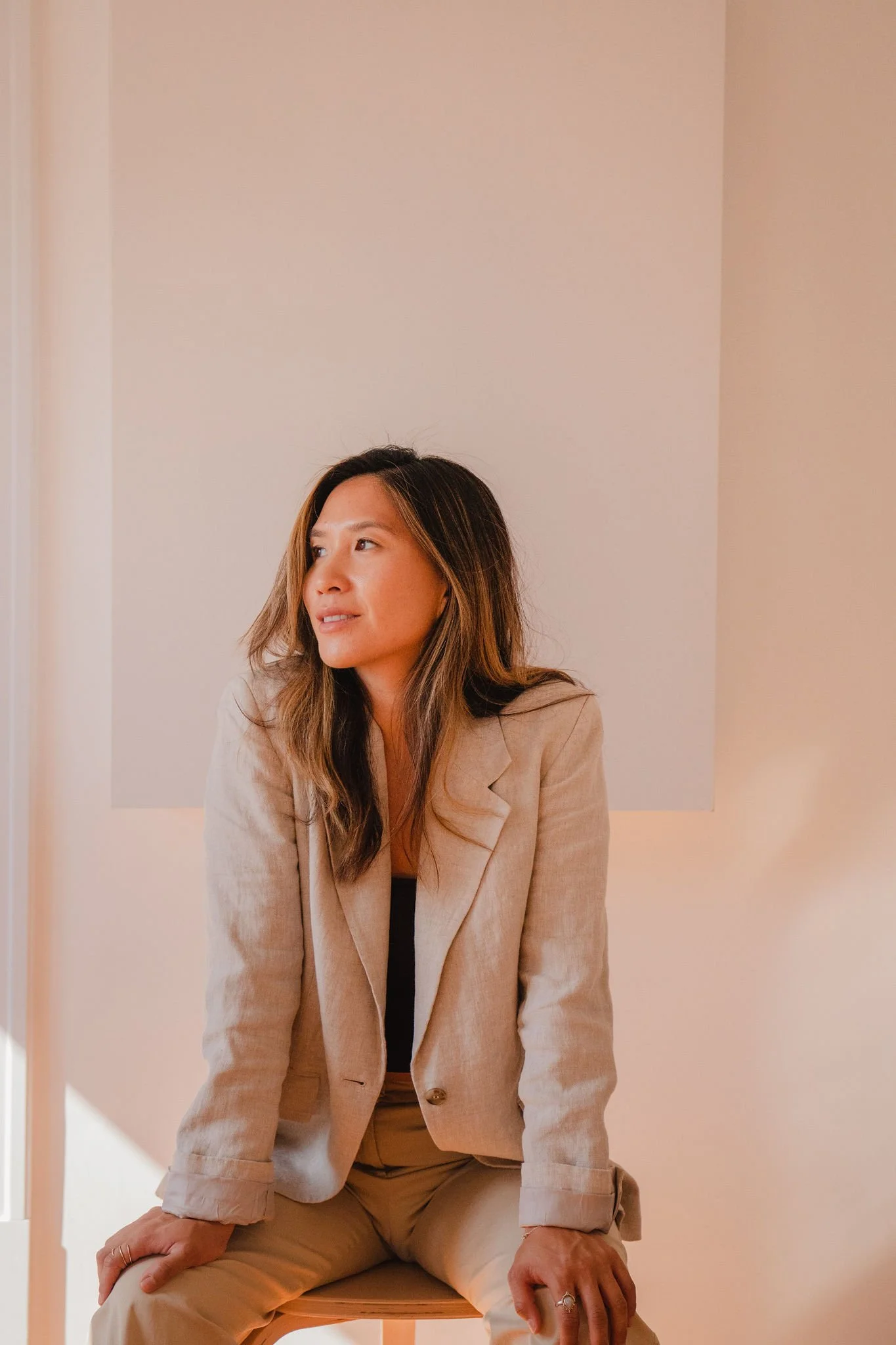 A woman with long brown hair sitting on a stool, wearing a beige blazer and pants, in front of a plain background.