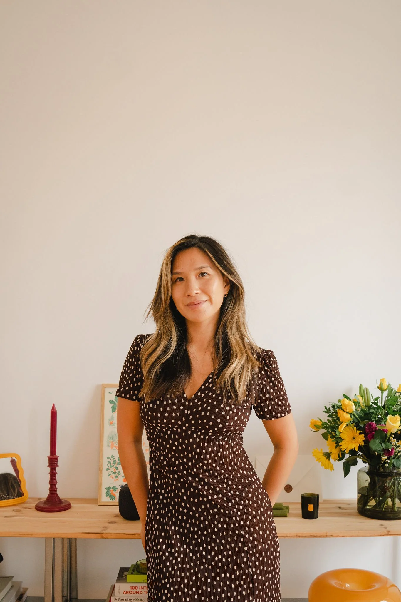A woman with wavy, shoulder-length hair, wearing a brown dress with white polka dots, standing in front of a light-colored wall, with a side table holding a red candle, a framed artwork, a black speaker, and a vase of yellow flowers.