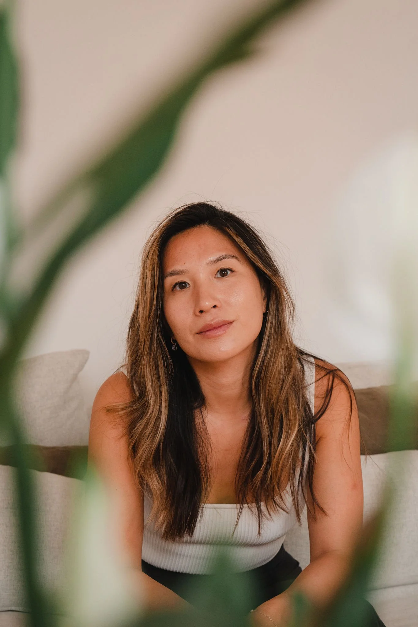 A woman with long wavy brown hair and light skin sits on a beige couch, looking at the camera, partially obscured by green plant leaves in the foreground.