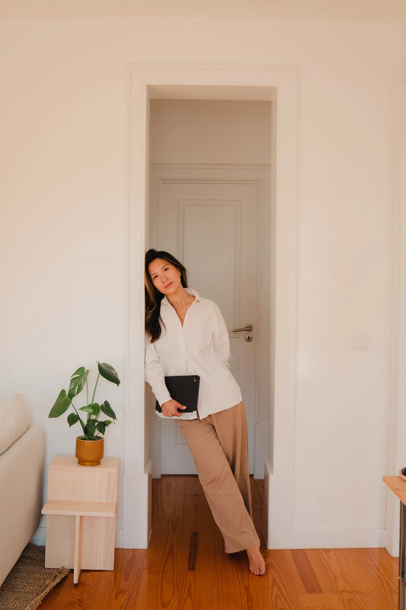 A woman with long dark hair and fair skin leaning against a doorframe in a minimalist room. She is wearing a white button-up shirt, beige pants, and is holding a black tablet or laptop. There is a small wooden side table with a potted plant on it to her left, and a beige sofa partially visible on the left side of the image.
