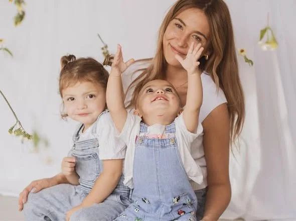 A woman with two young girls, one sitting on her lap and the other reaching up to touch her face, in a bright room with plants and white curtains.