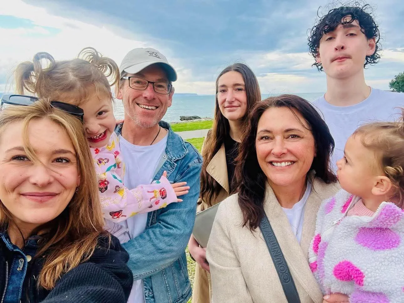 A group of seven people, including two young girls, two women, two men, and a teenager, smiling and posing for a photo outdoors near a body of water with a cloudy sky.