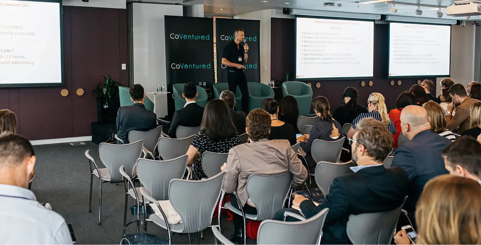 A professional conference with a speaker presenting to an audience in a modern room. The speaker stands on a stage, holding a microphone, with presentation slides displayed on large screens behind him. The audience is seated in rows, listening attentively.