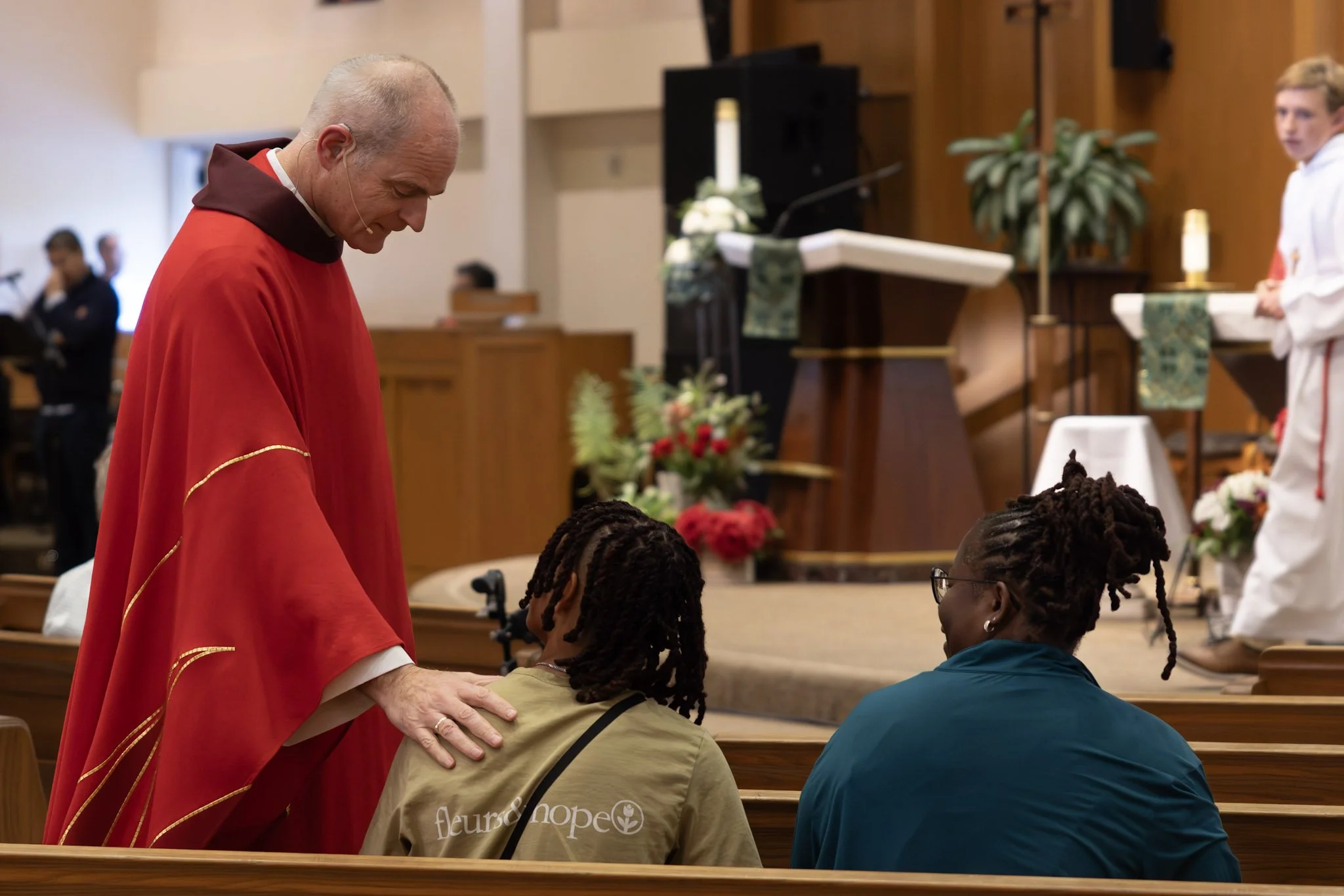 A priest in red robes comforting a woman and a girl sitting in a church pew during a service