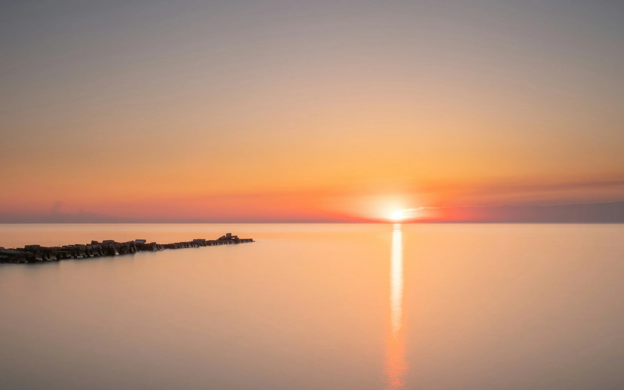 Sunset over calm water with a narrow pier on the left side extending into the water, and colorful sky with shades of orange, pink, and purple.