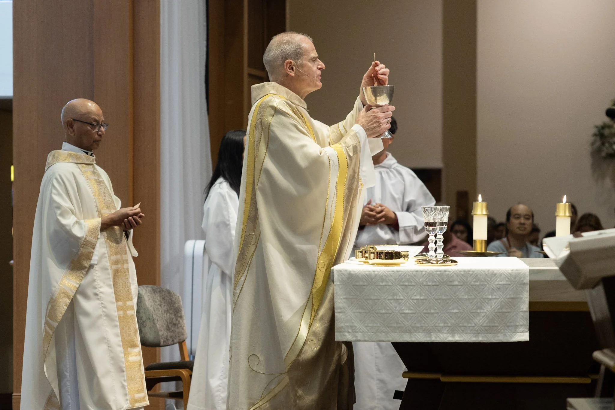 A Catholic priest and altar servers perform the Eucharist during a religious service inside a church, with candles and chalice on the altar.