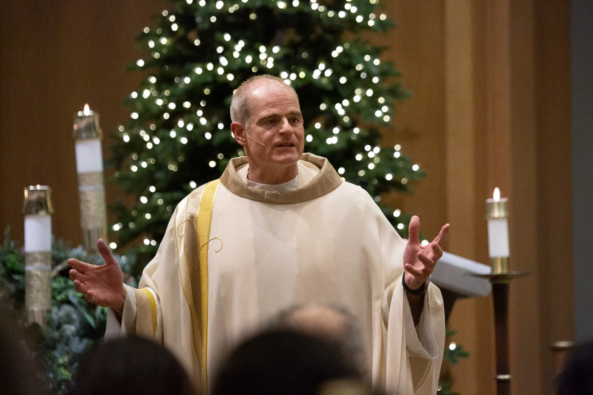 A priest stands in front of a lit Christmas tree with white candles on stands on either side, speaking to an audience in a church.