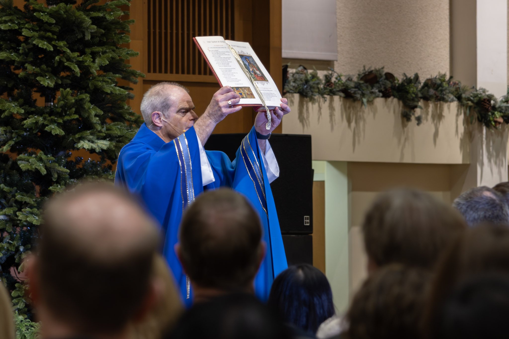 A church minister wearing a blue robe holds an open book above his head during a religious service, with congregation members watching.