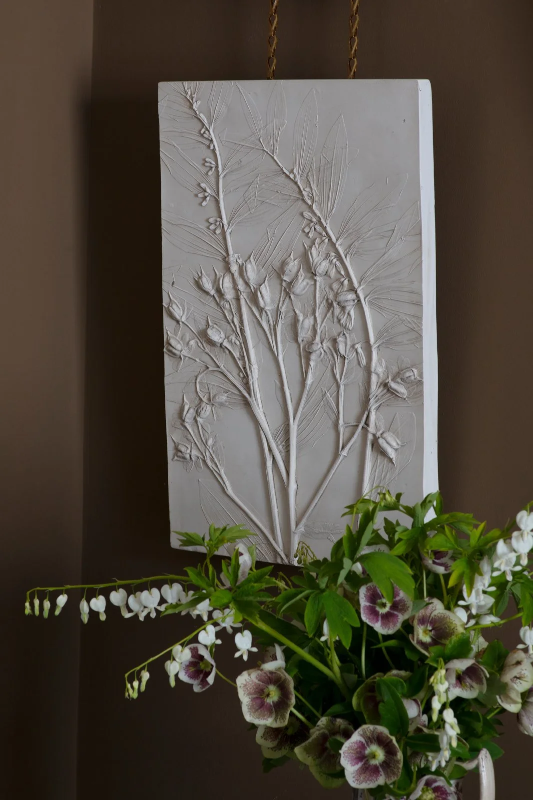 An unpainted plaster cast with embossed botanical design, placed above a vase of white and purple flowers.