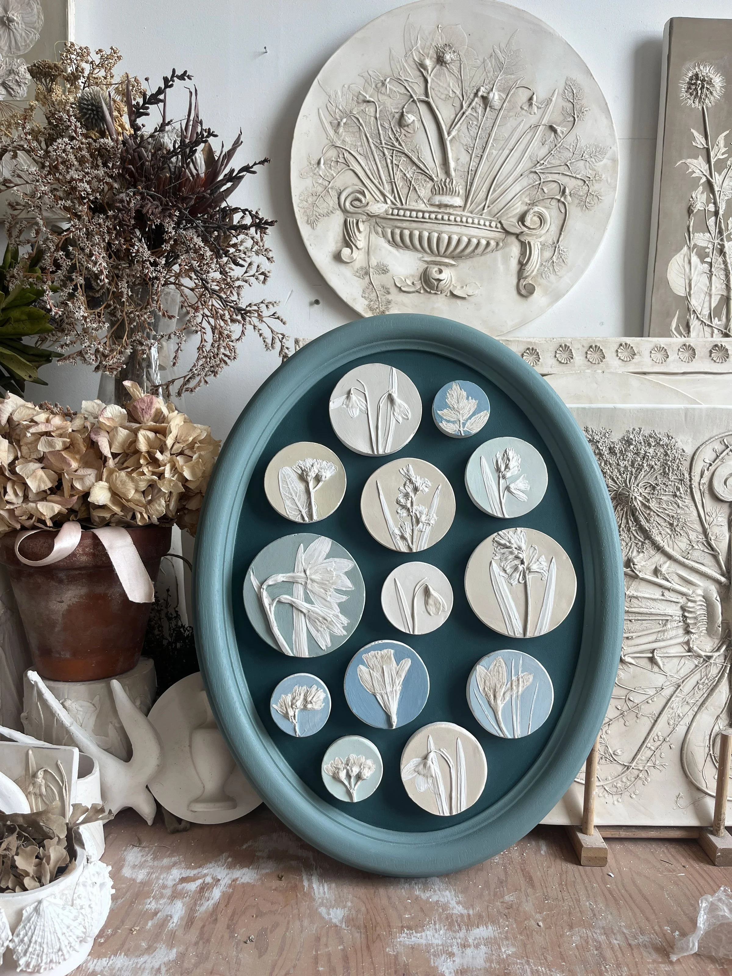 A blue oval frame containing smaller circular relief artworks of botanical flowers and leaves in white, set against a teal background. Surrounding the frame are dried flowers and other botanical relief artwork pieces displayed against a white wall.