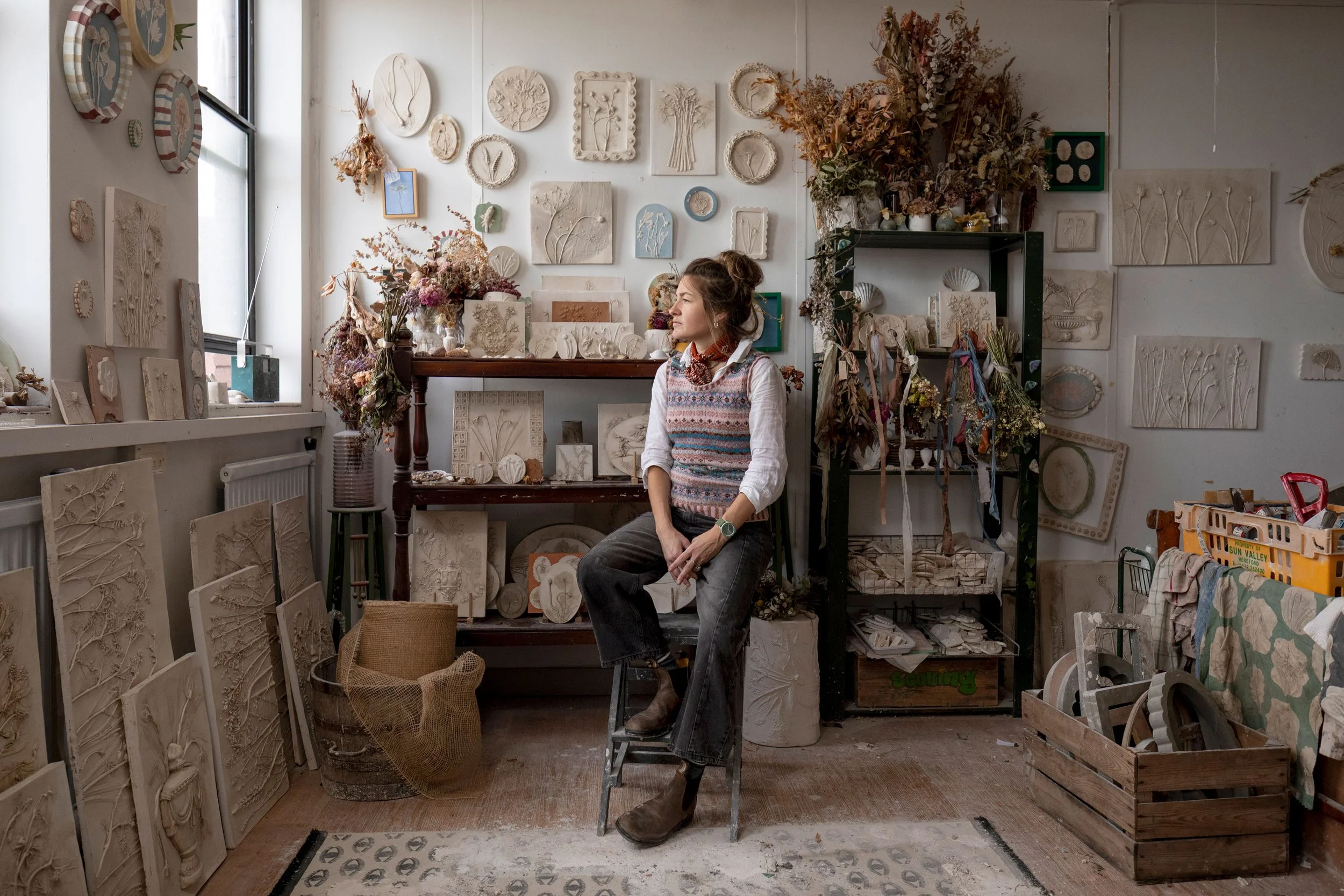Katy Eccles sitting on a stool in a studio surrounded by various art pieces, dried flowers, and art supplies, with walls decorated with textured relief artworks and ceramics.