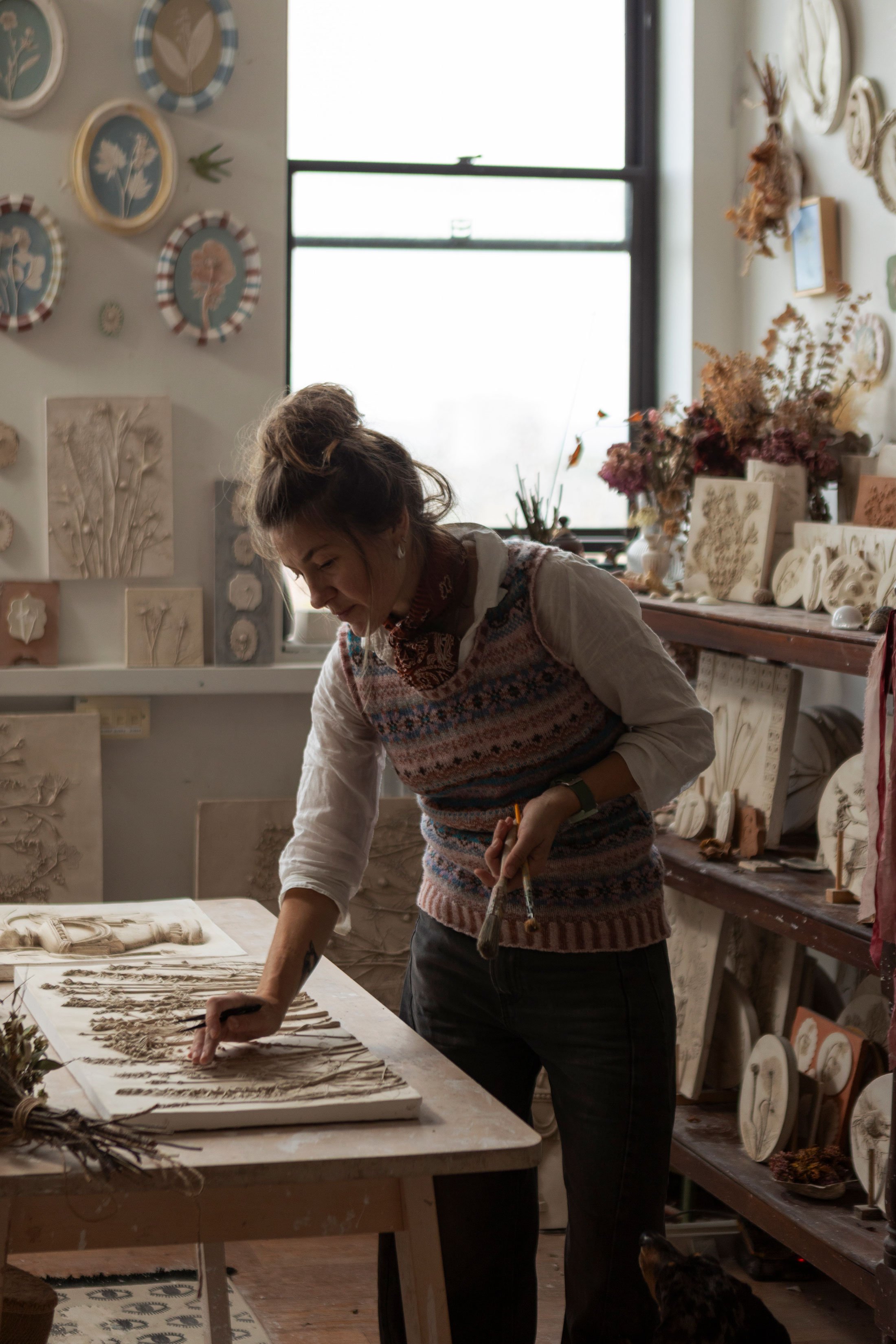 Katy Eccles in her studio working on a botanical plaster cast with shelves of finished pieces and wall art.