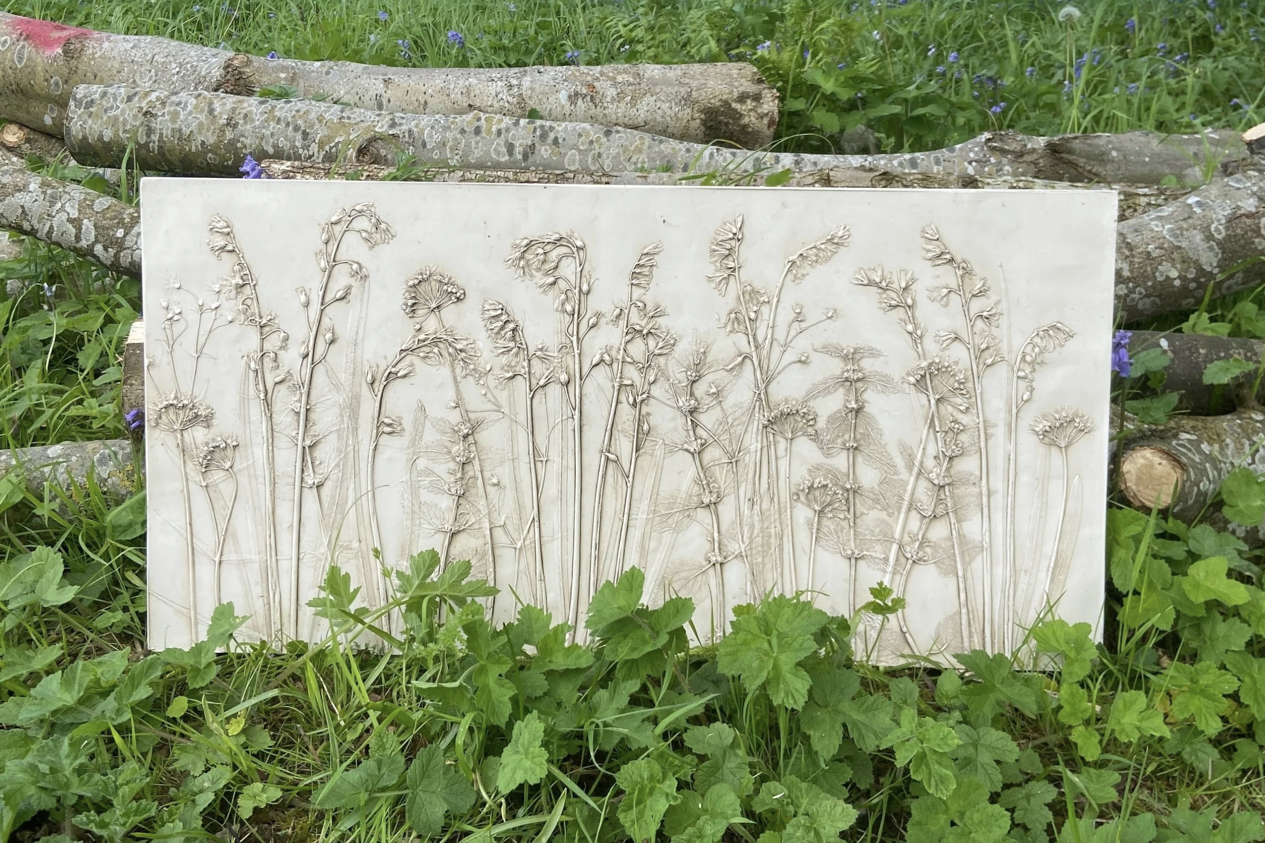 A plaster cast with embossed wildflowers design placed on the ground amidst grass and green plants, with logs and purple flowers in the background.