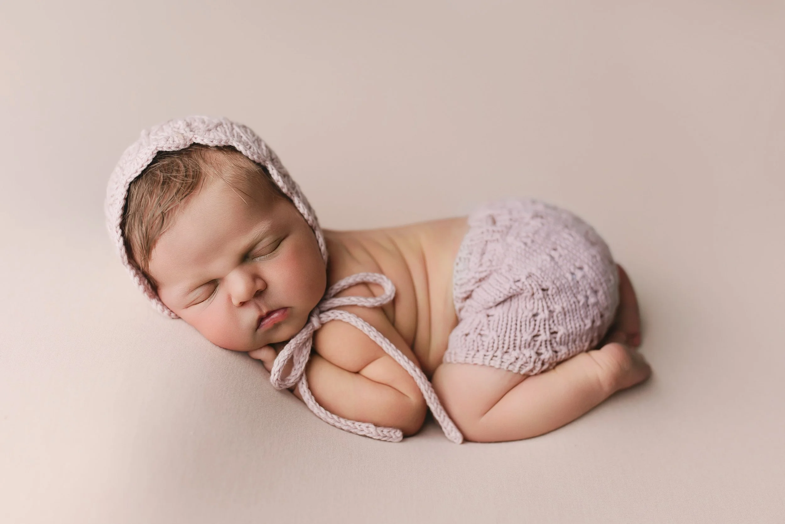 Søvnende babyjente med lyserosa lue og Shorts liggende på mage på en beige bakgrunn.