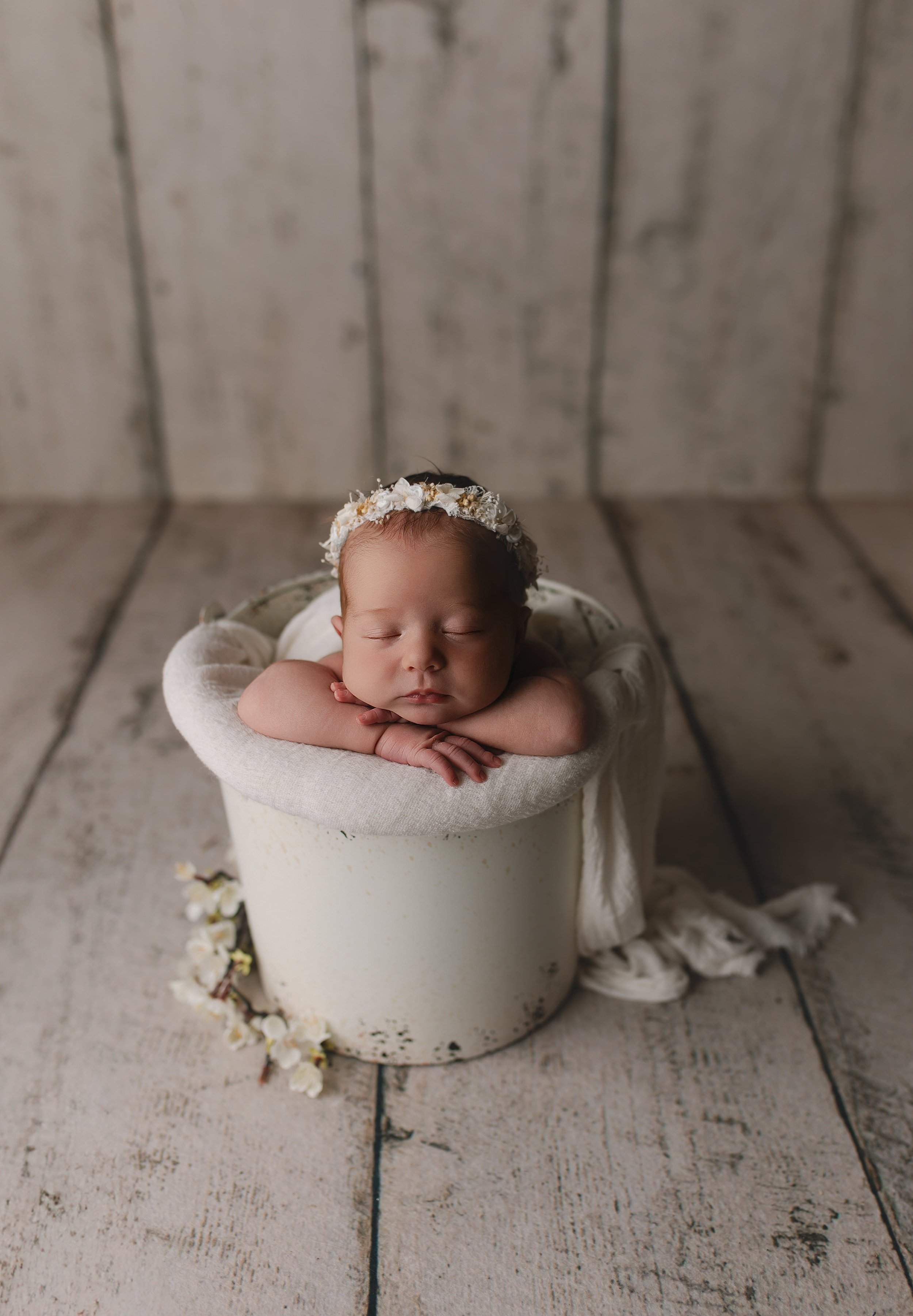 Søvnende baby med blomsterkrans på hodet, hvilende på en pute i en rund kurv med trekanter på og lyst stoff rundt. Bakgrunnen er et trepanel i lyse farger.