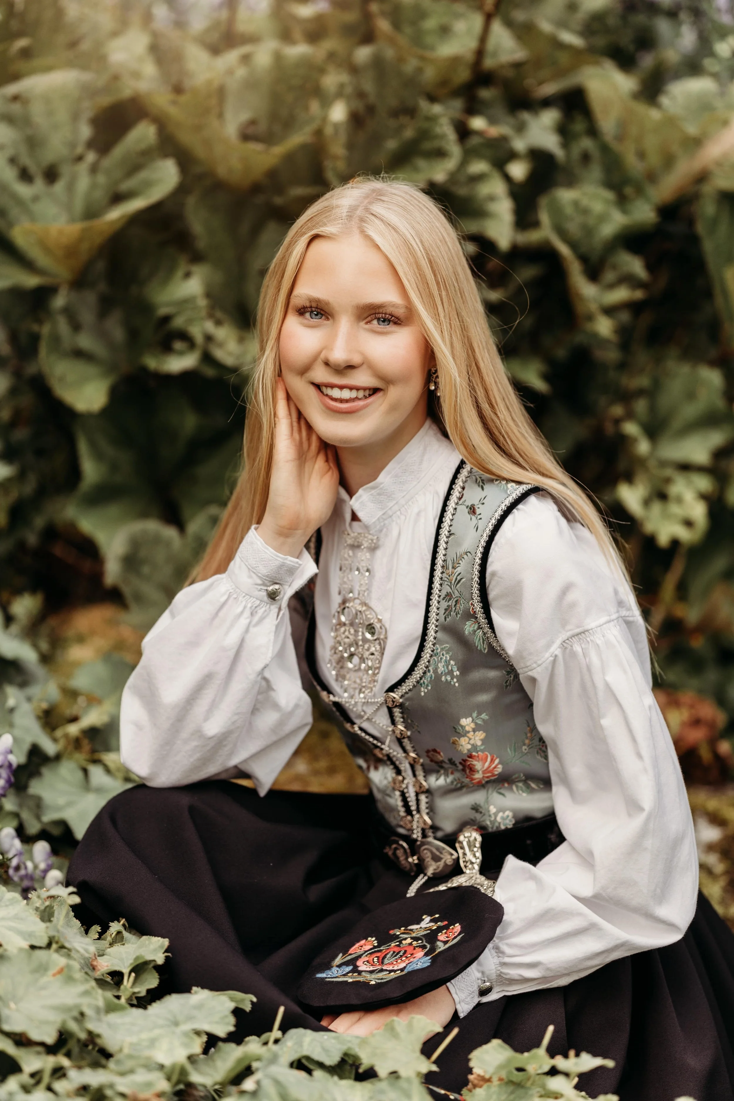 En ung kvinne med langt blondt hår, iført tradisjonell norsk bunad, smiler og sitter i en hageseng med grønne planter rundt.
