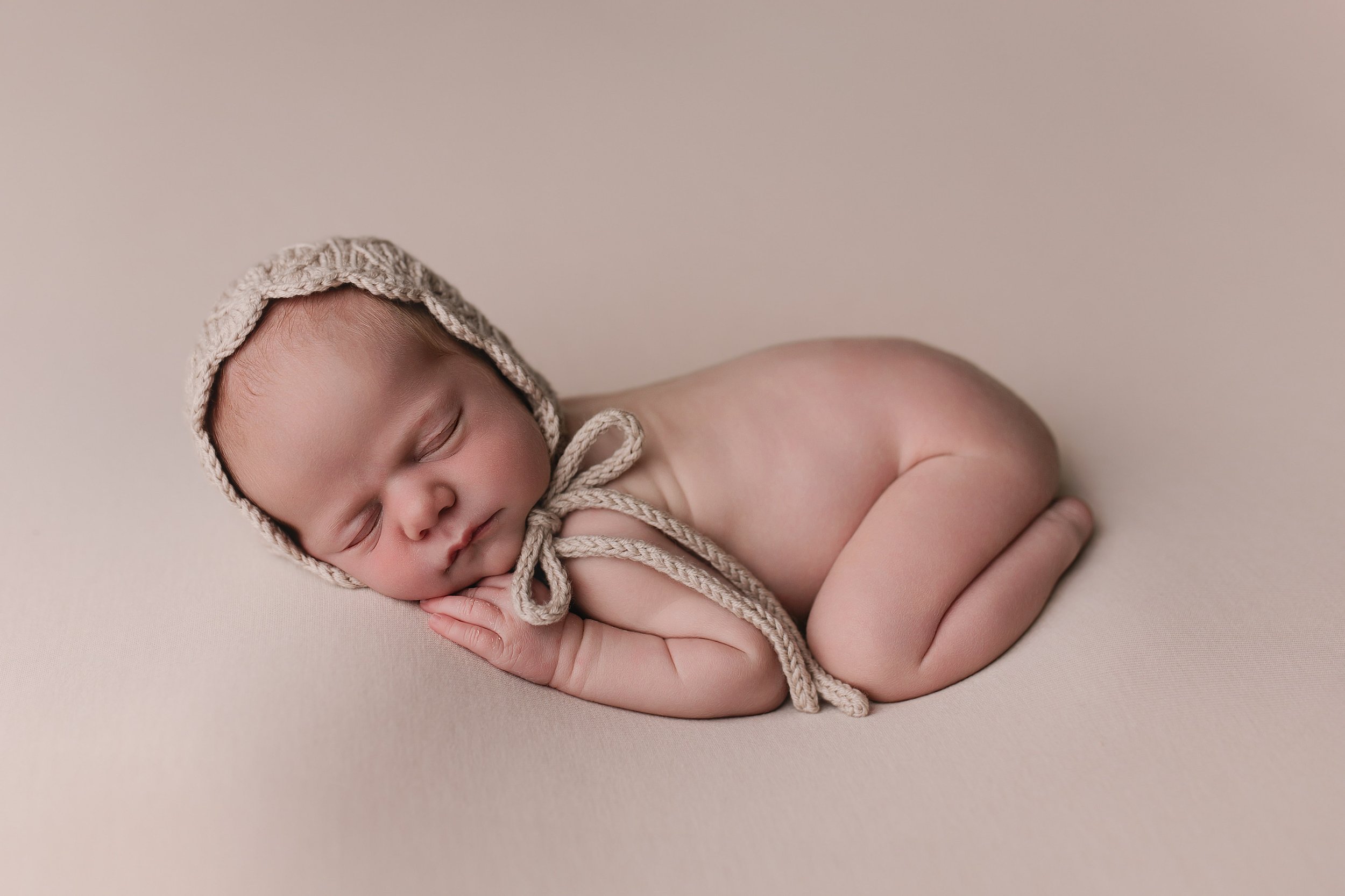 Søvnende baby med bølget beklædningshue, liggende på siden på en beige bakgrunn, med hender under ansiktet.