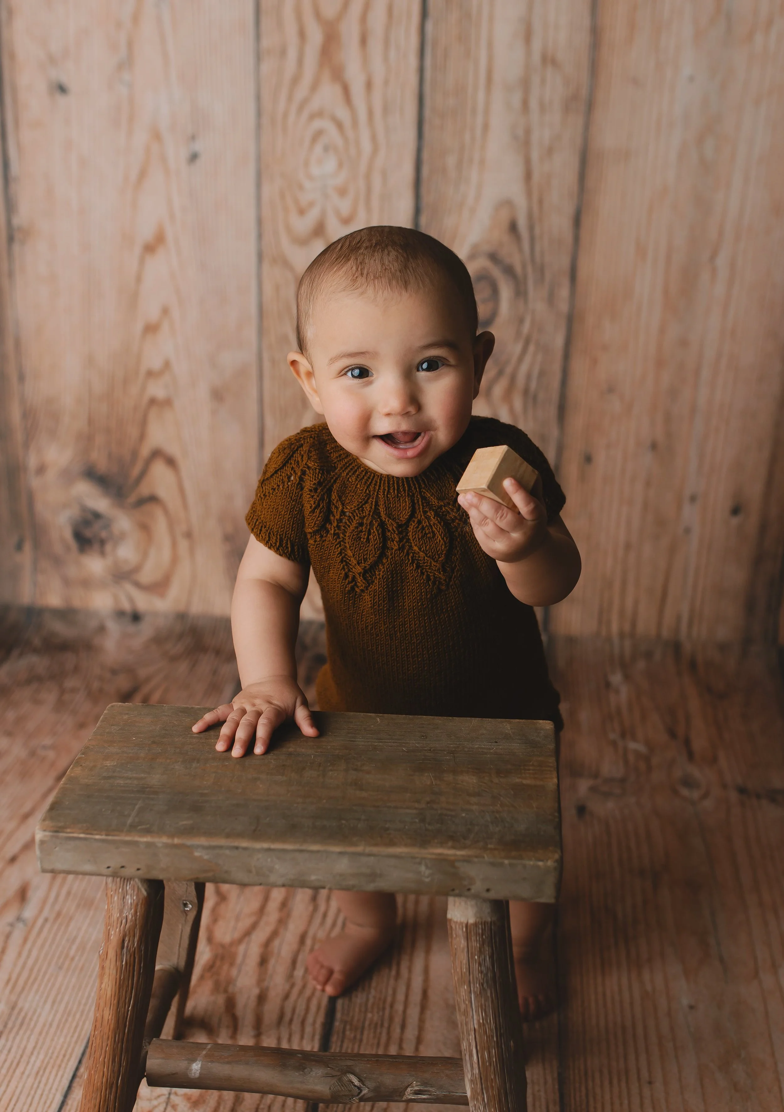 Liten gutt med kort hår som står bak en liten trestol, holder en blokk i hånden og smiler mot kamera. Bakgrunnen er trepanel.