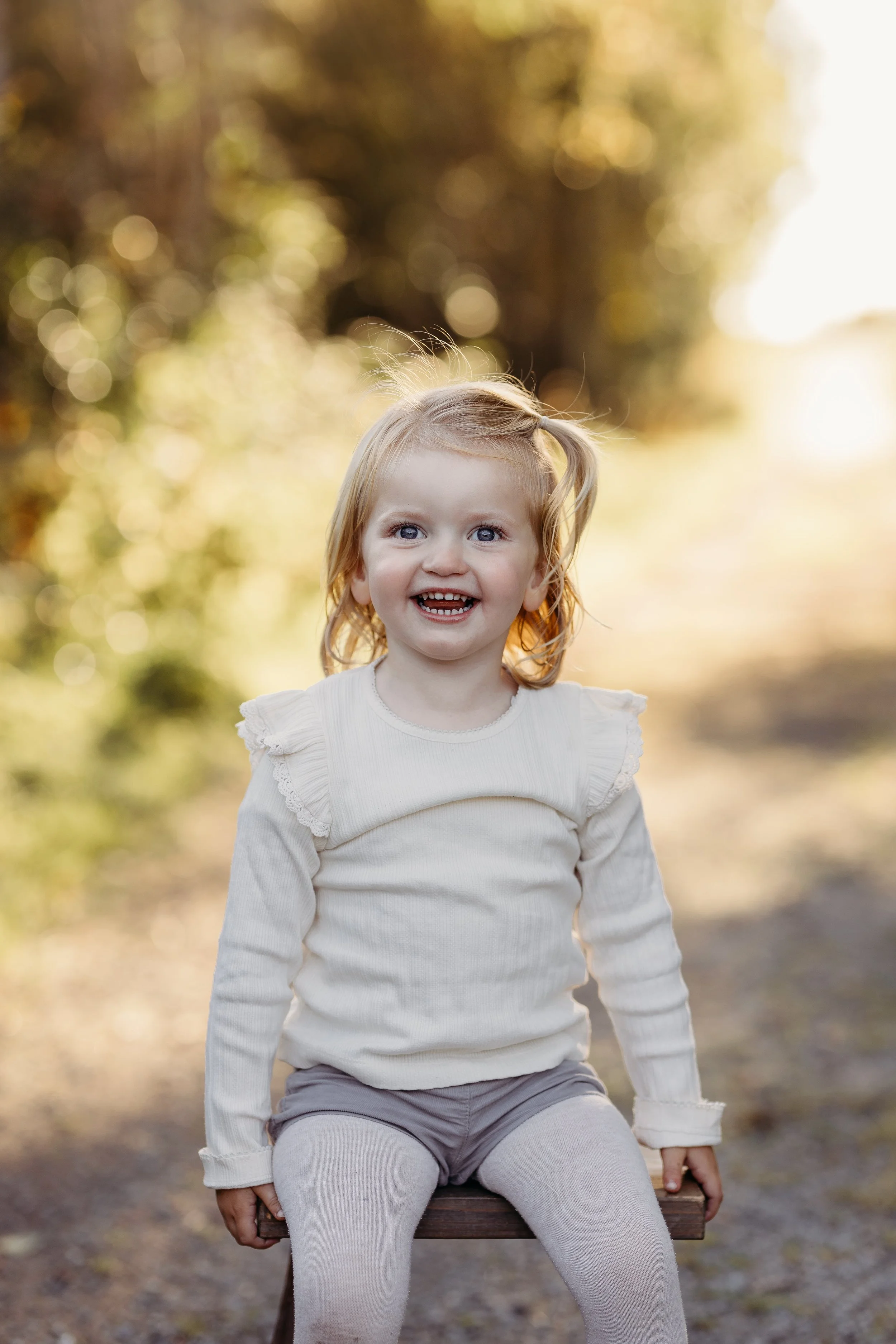 Smilende liten jente med lyst hår sitter på en benk utendørs om høsten, omgitt av trær med gult løv.