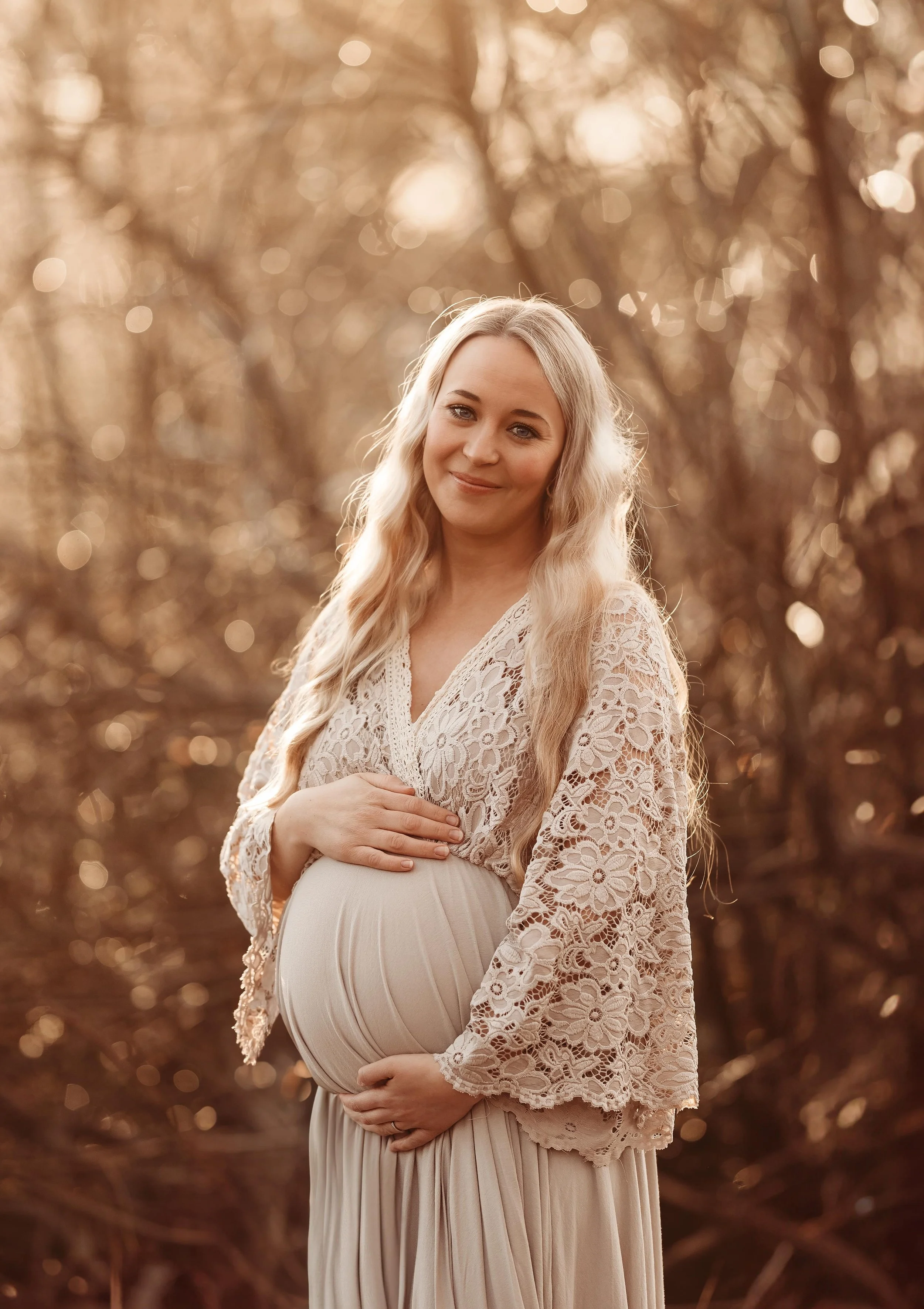 En gravid kvinne står i en skog med gylden belysning, iført en hvit blondekjole og smiler mildt.
