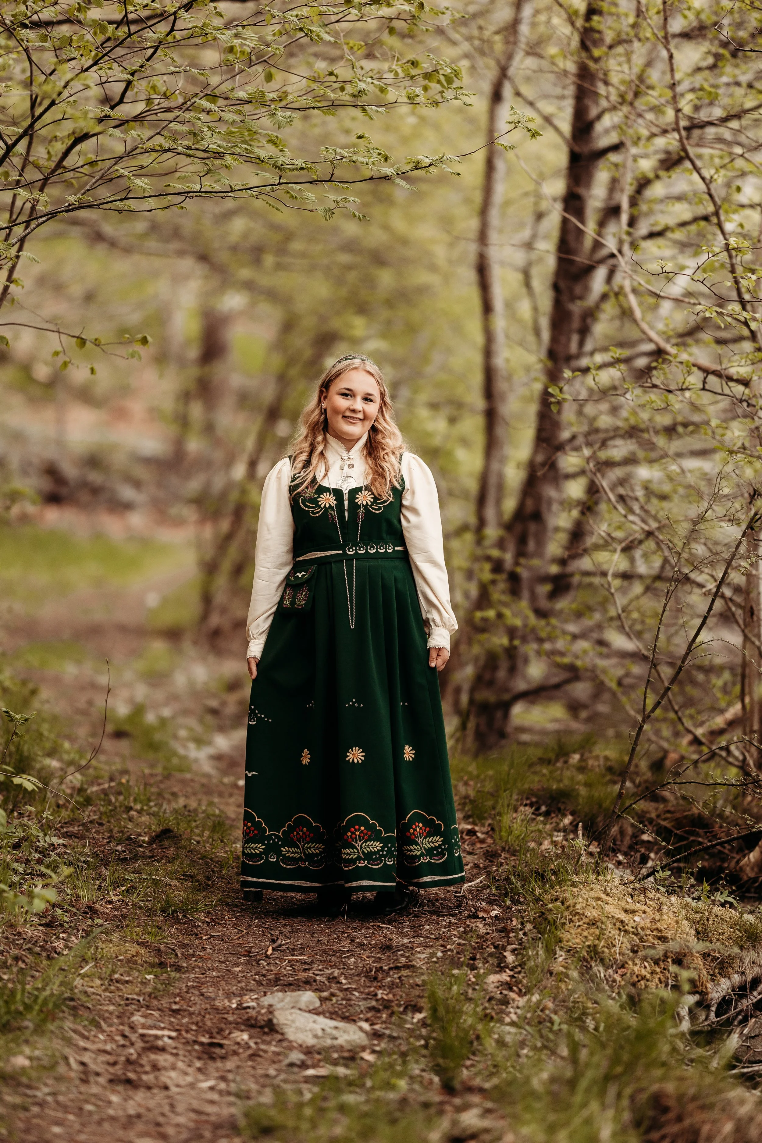 En jente ikledd tradisjonell norsk bunad står på en skogssti med grønne blader og trær rundt.