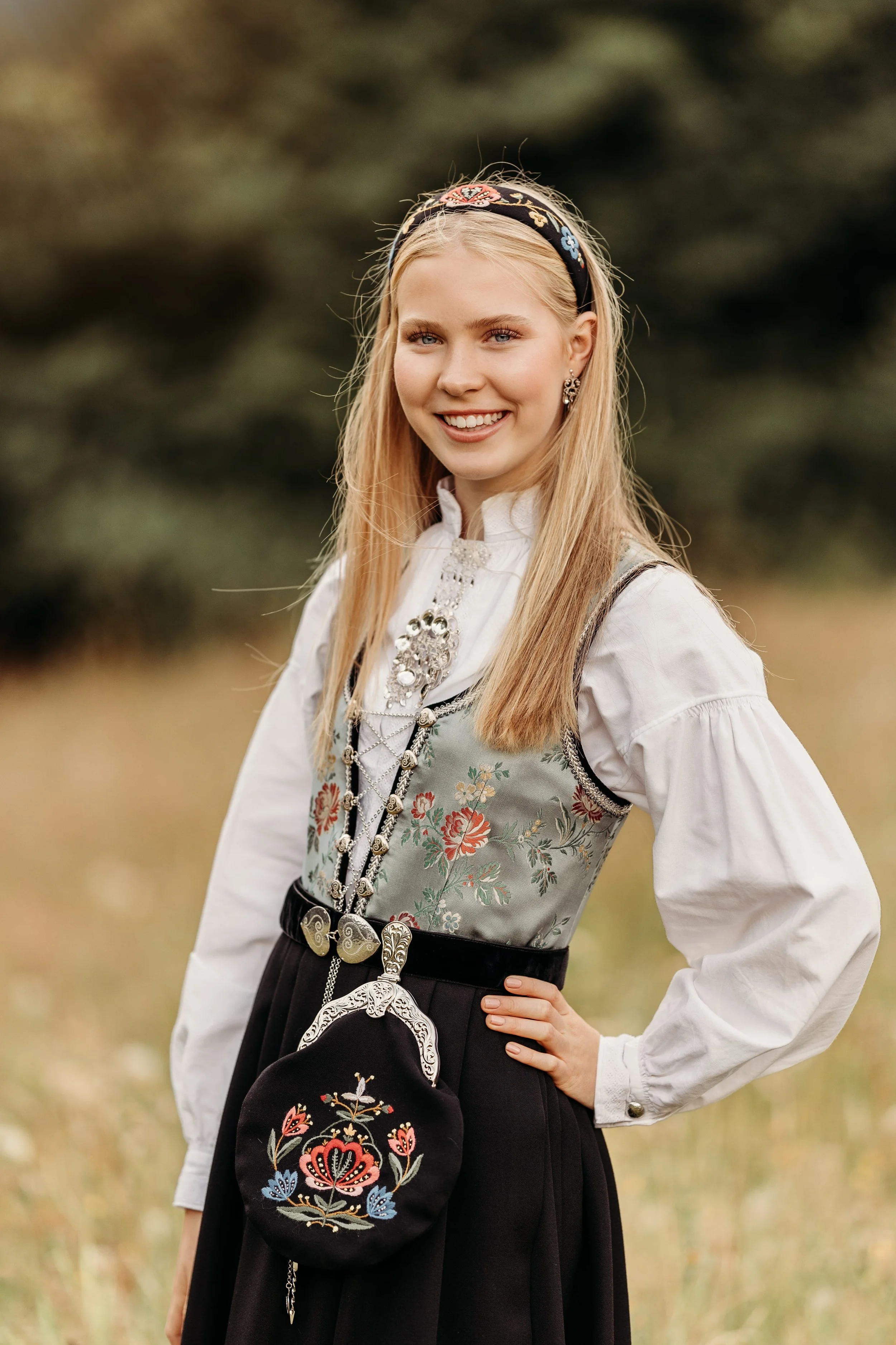 En ung kvinne i tradisjonell norsk bunad med blomstrete vest, hvit skjorte, og et bånd med broderier over skulderen, står i en utendørs natur med grønt gress og trær i bakgrunnen.