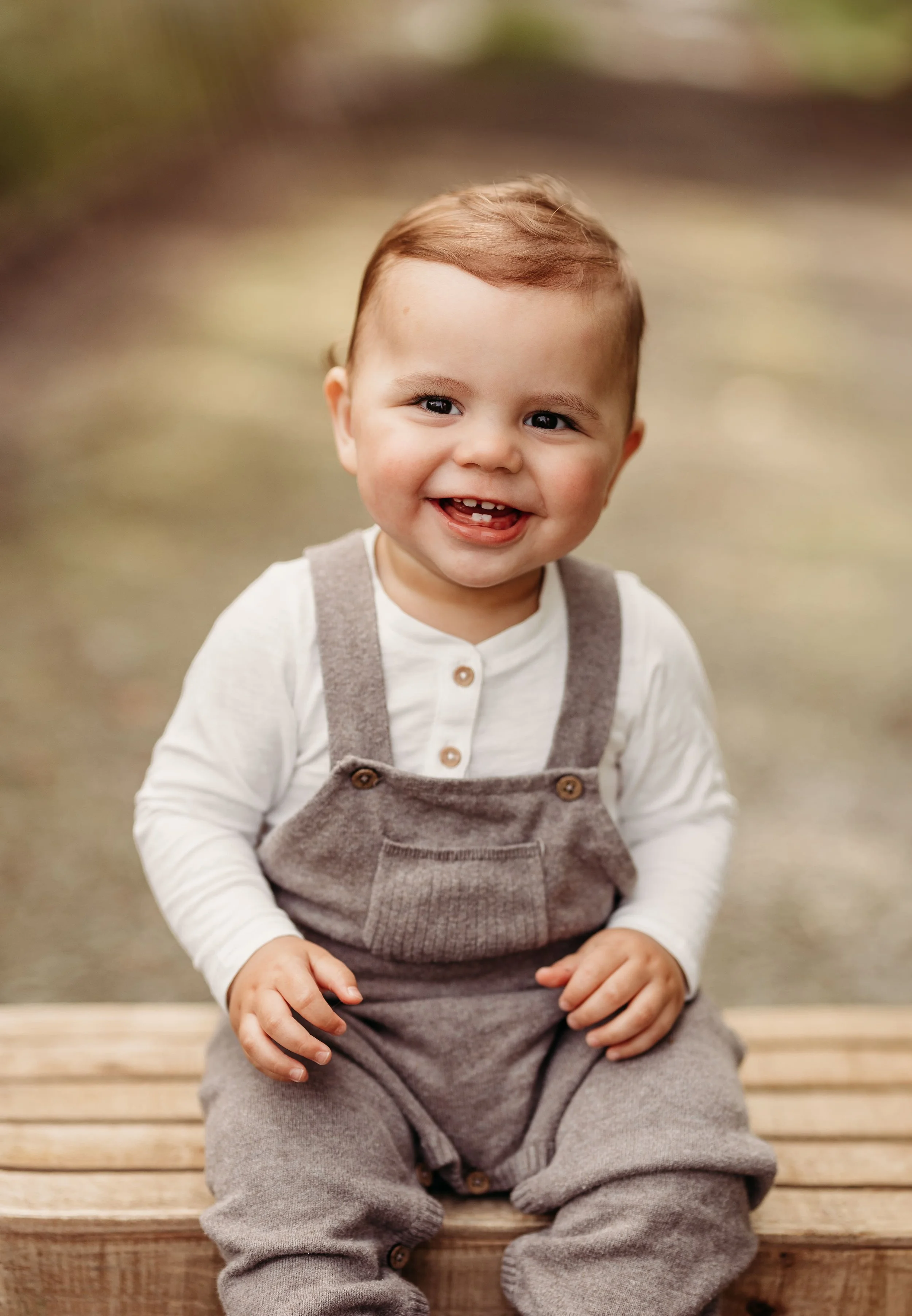En smilende liten gutt med lyst hår, som sitter på en trebrygge, iført en hvit langermet skjorte og gråt overalls. Bakgrunnen er uklar, og det ser ut til å være utendørs.