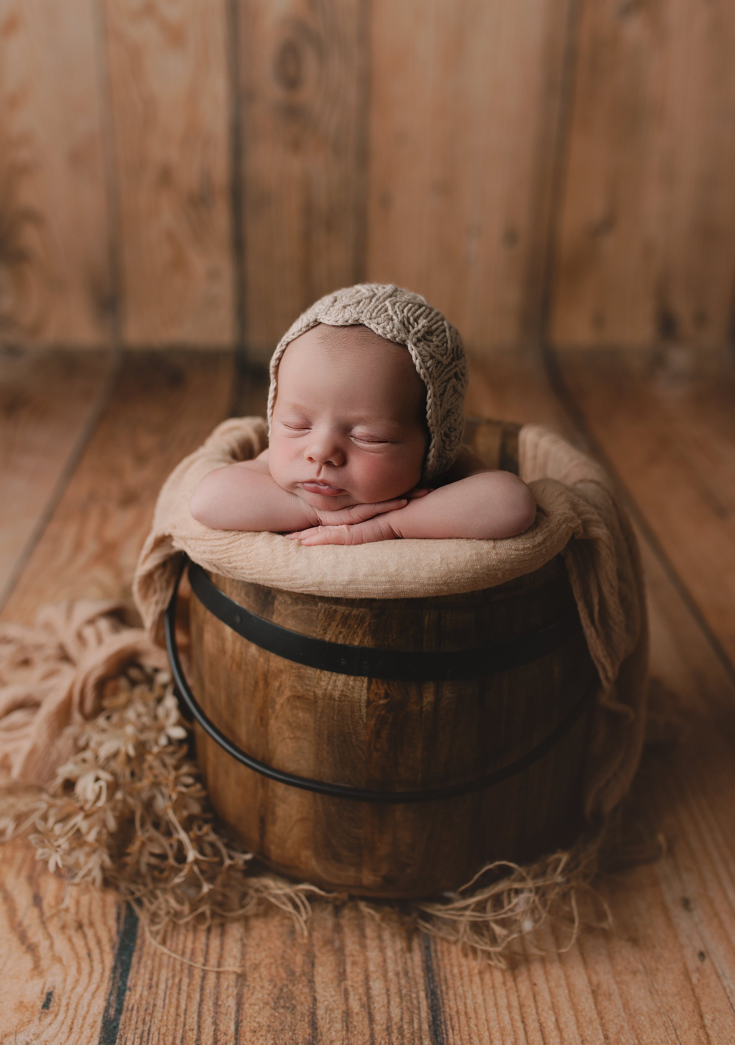 Søvnende baby med lue, liggende i en rund mahogni-tønde, omgitt av et beige stoff og tørket blomster på en trebunn.