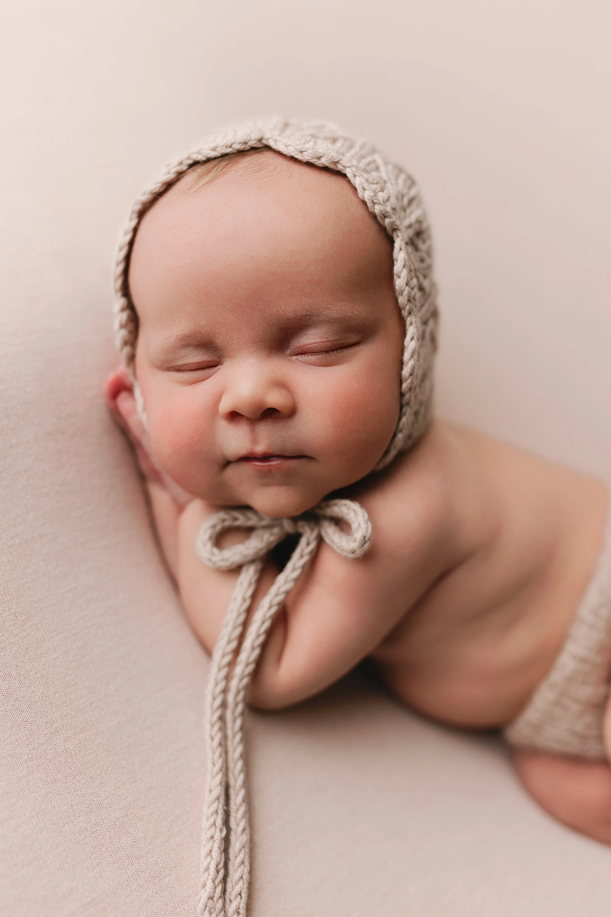 Søvnende baby med knapp av ullhatt, liggende på lyse stoff, med lukkede øyne og rolig uttrykk.