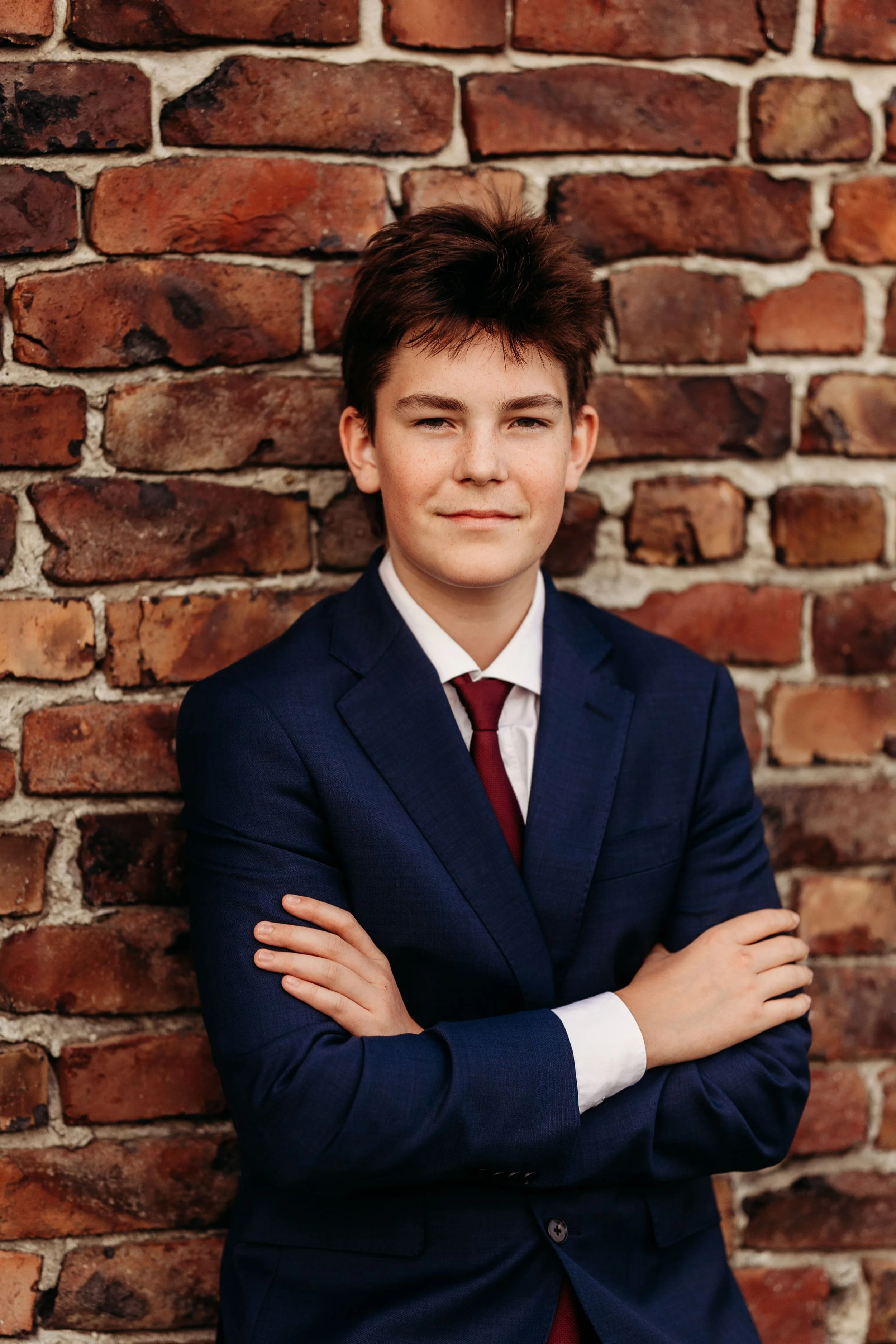 En ungdom mann står foran en mur av murstein, kledd i blå dress, hvit skjorte og rød sløyfe, med armene krysset.