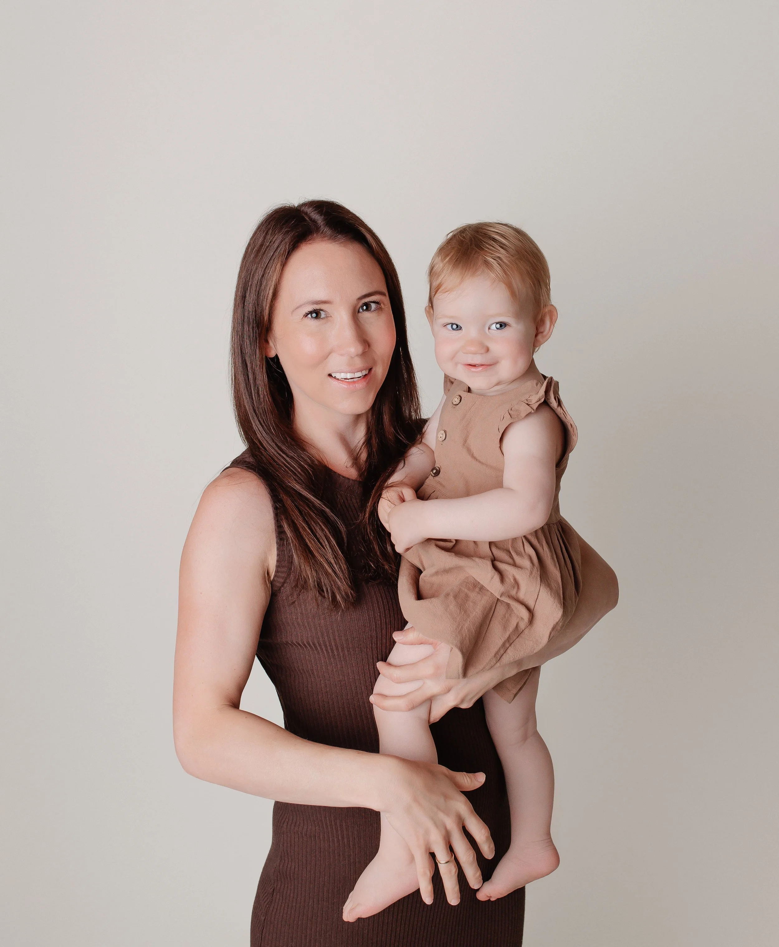 En kvinne med langt brunt hår holder en liten barn med lyst hår i armene, begge smiler mot kameraet, mot en enkel lys bakgrunn.