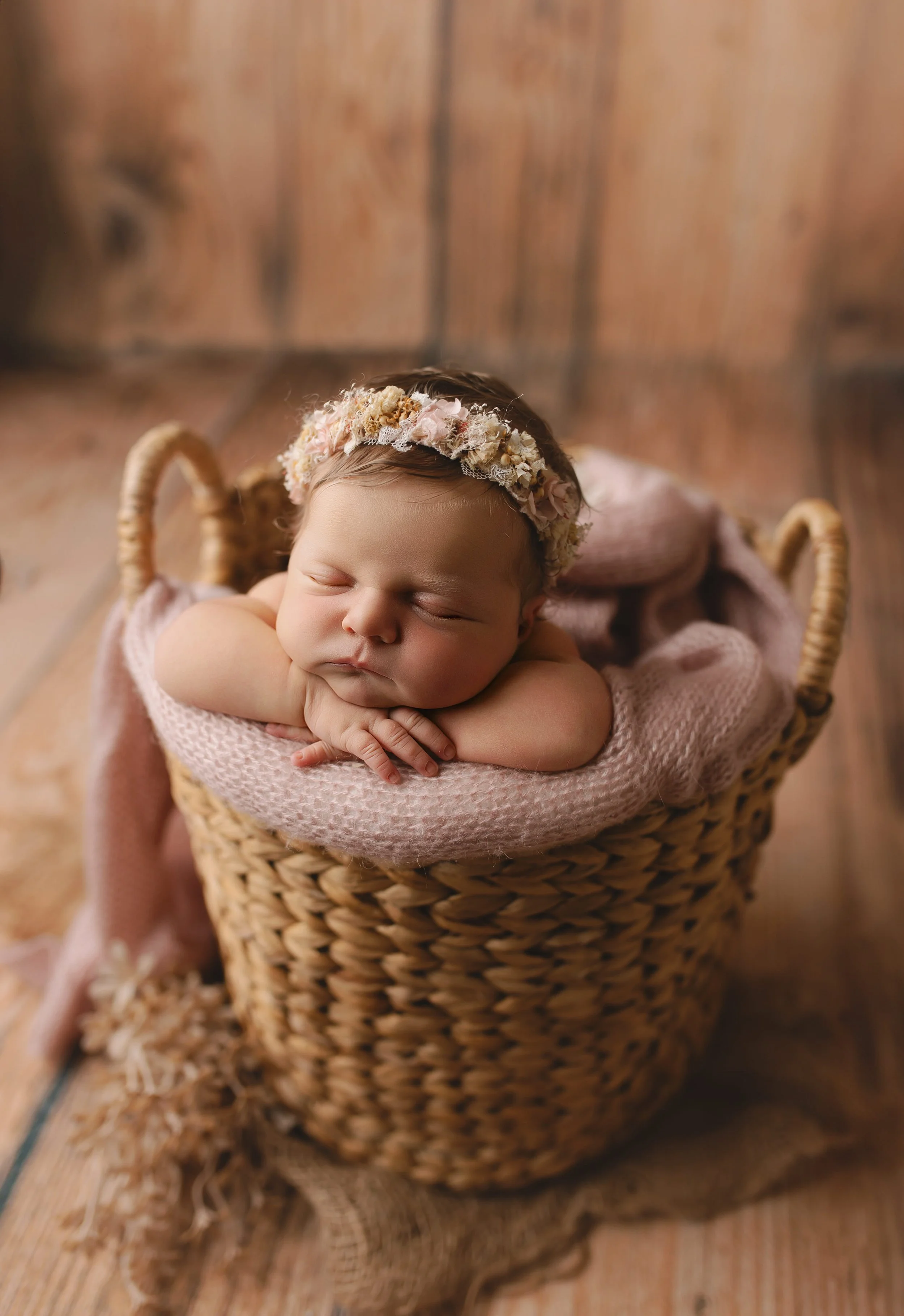 Søvnende baby med blomsterkrans i håret, plassert i en kurv med en rosa ullpledd, på en treplate.