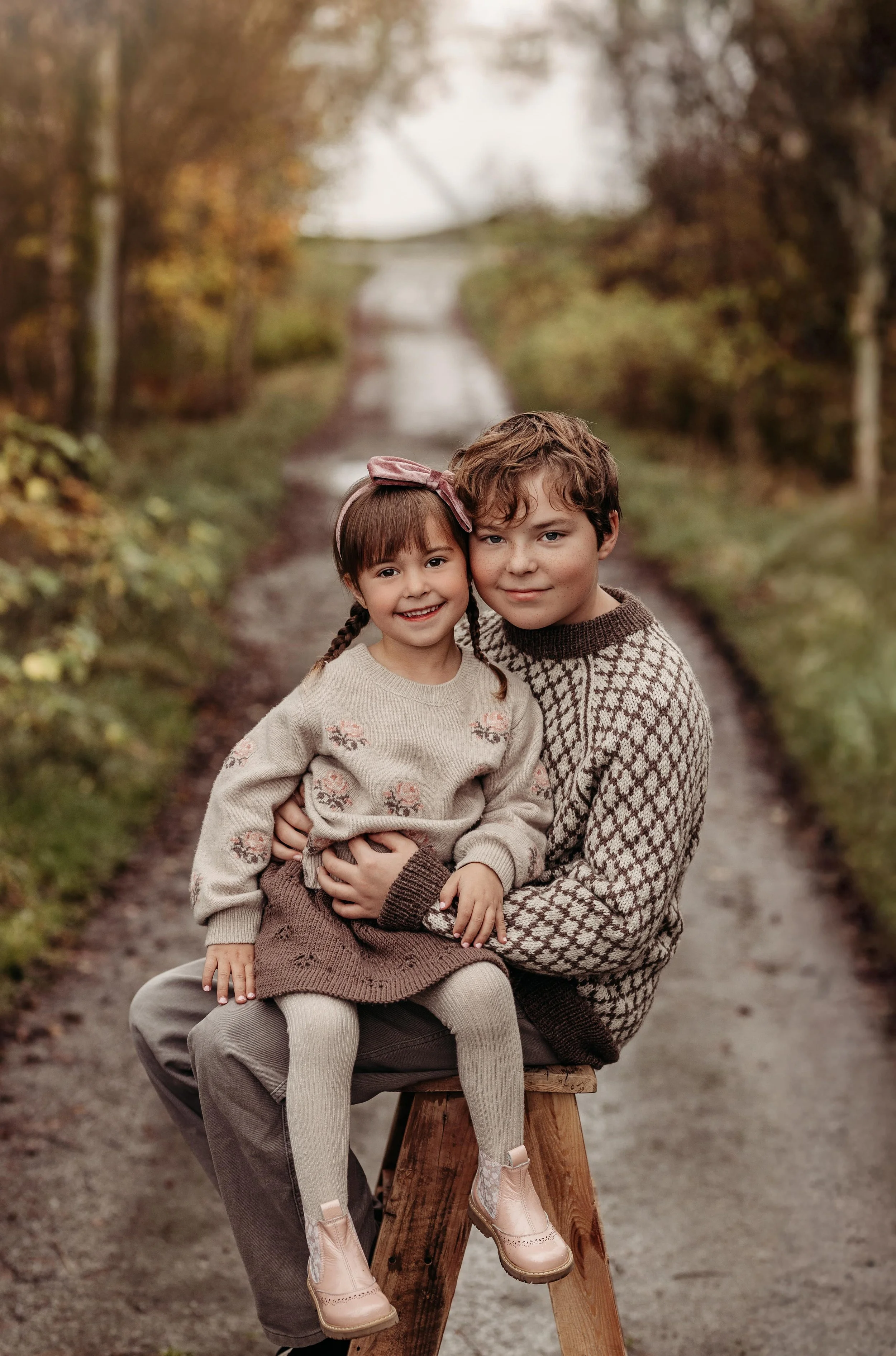 To barn, en jente med brunt hår og en gutt med krøllete hår, sitter sammen på en trebenk på en grusvei omgitt av trær med høstløv.