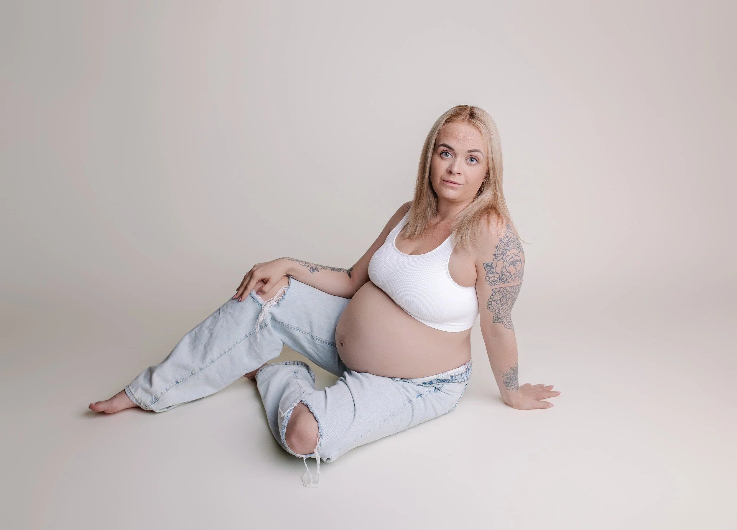 En gravid kvinne sitter på gulvet mot en lys bakgrunn, iført hvit sports-BH og lyse, revne jeans.