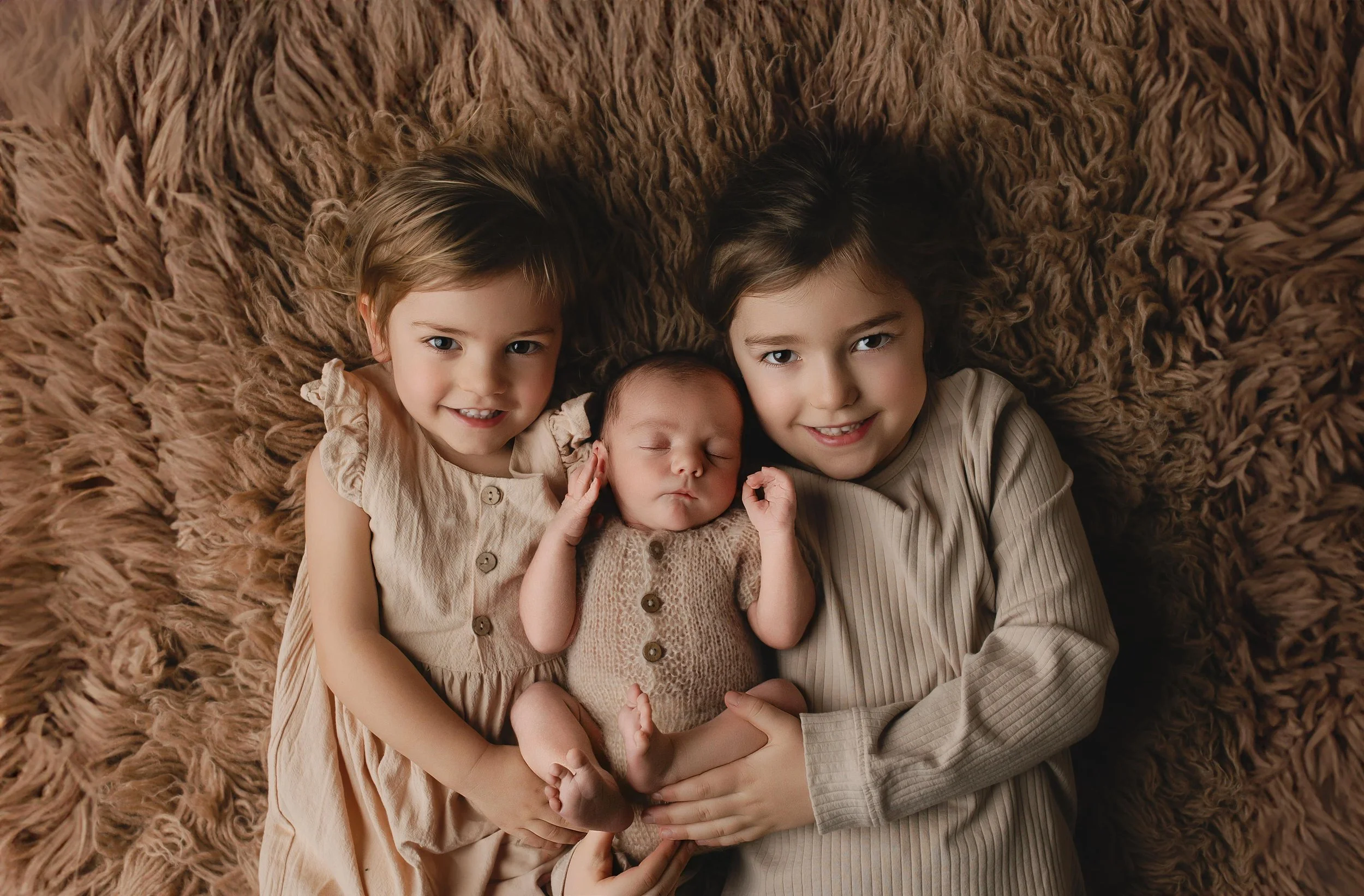 Tre små barn, to jenter og en baby, ligger på en myk, brun pels, smiler og ser mot kamera.