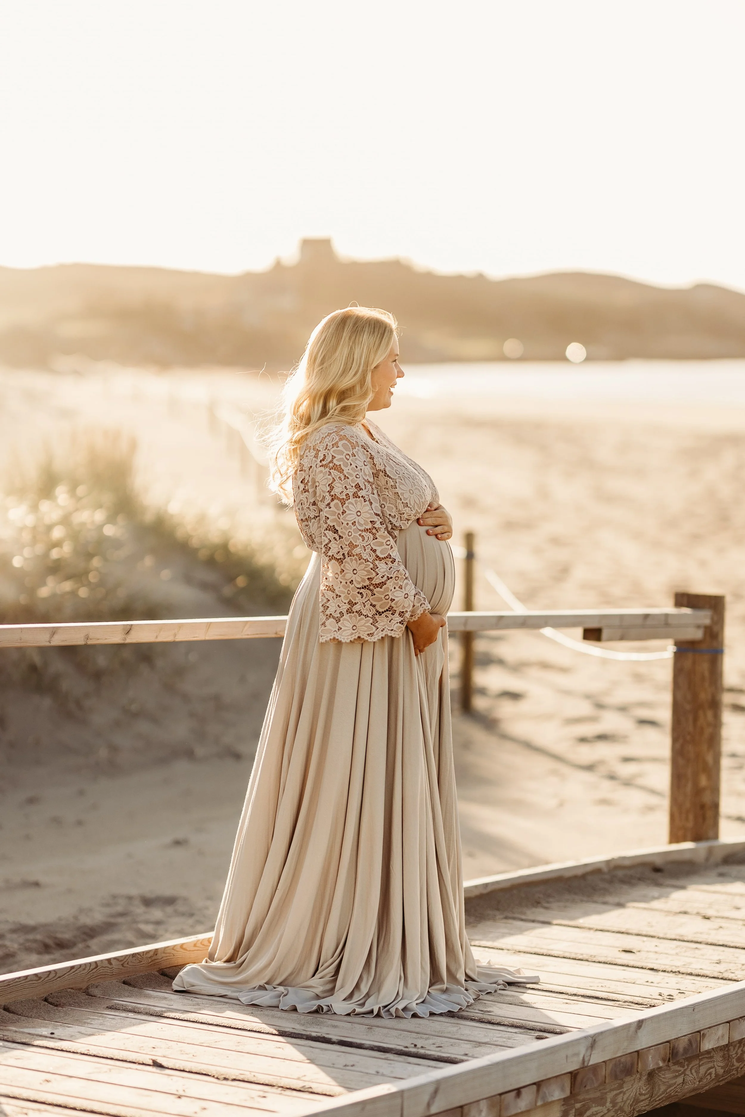 En gravid kvinne står på en treplattform ved stranden, kledd i en lang, hvit kjole med blondeyoverdel, under solnedgang.