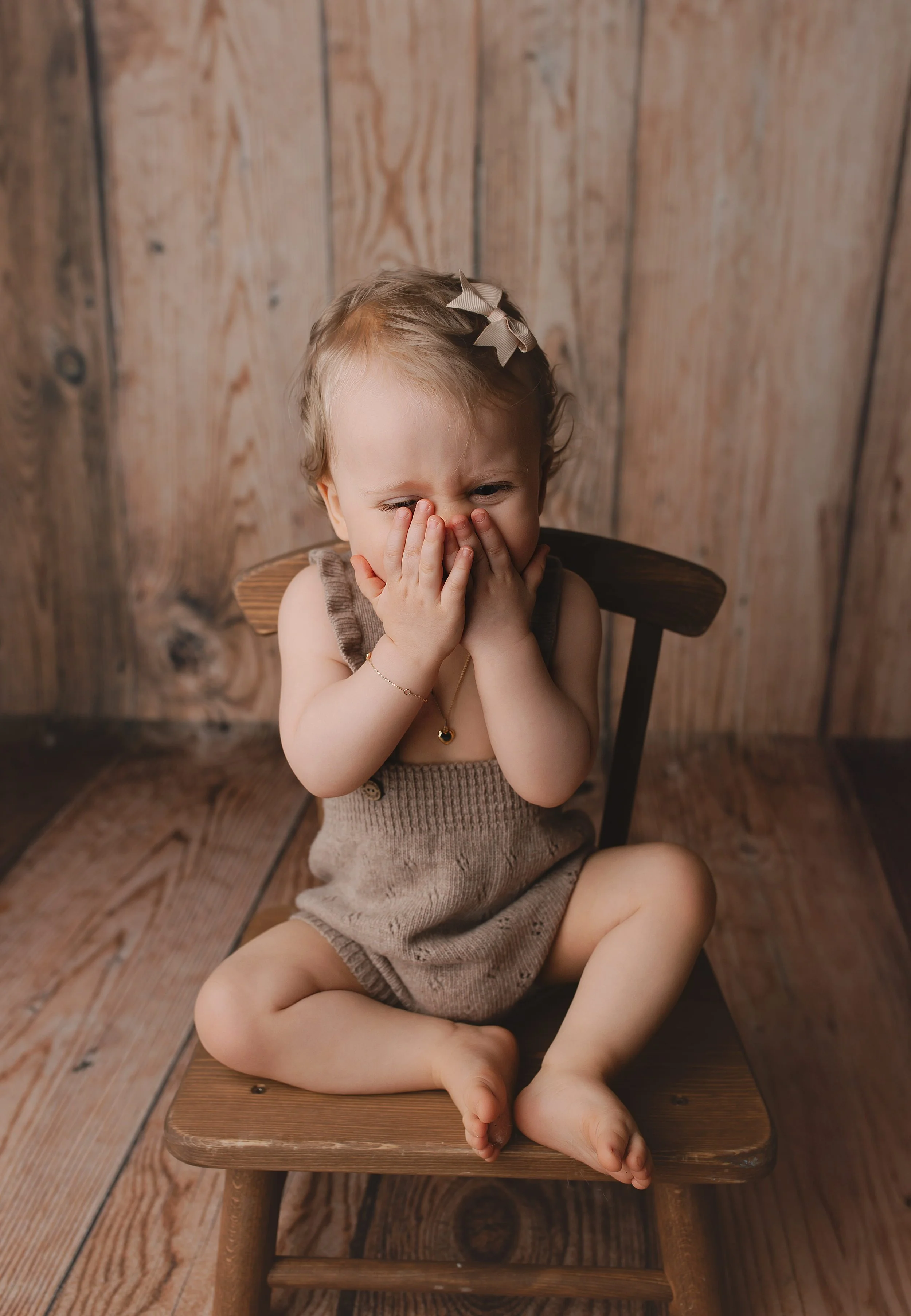 Liten jente sitter på en liten trebænk med hendene foran ansiktet, har et tau rundt halsen, og er iført en beige overalls og hårbånd med sløyfer, mot en trebakgrunn.