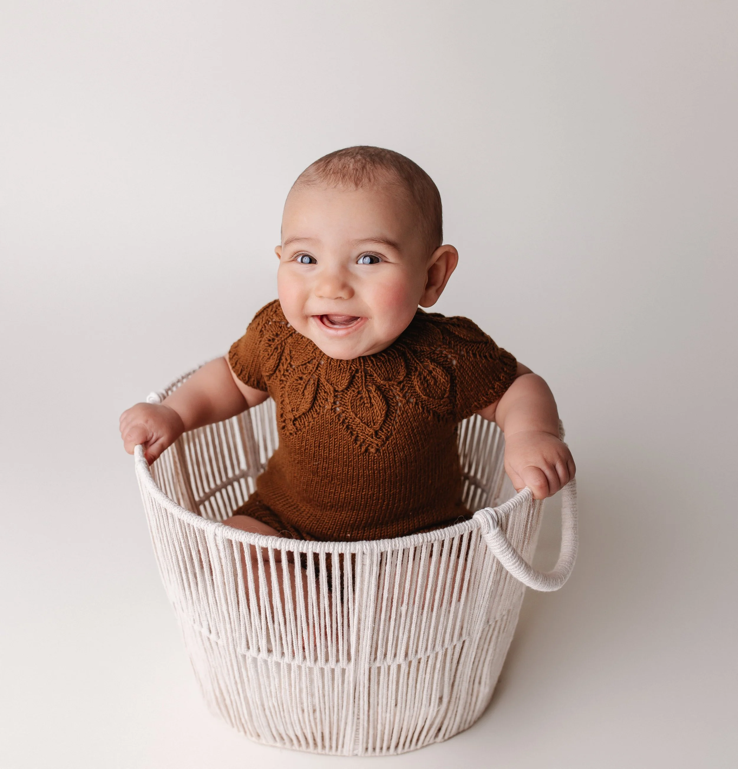 En glad baby med kort hår som sitter i en hvit kurv iført en brun strikkegenser. Bakgrunnen er hvit.