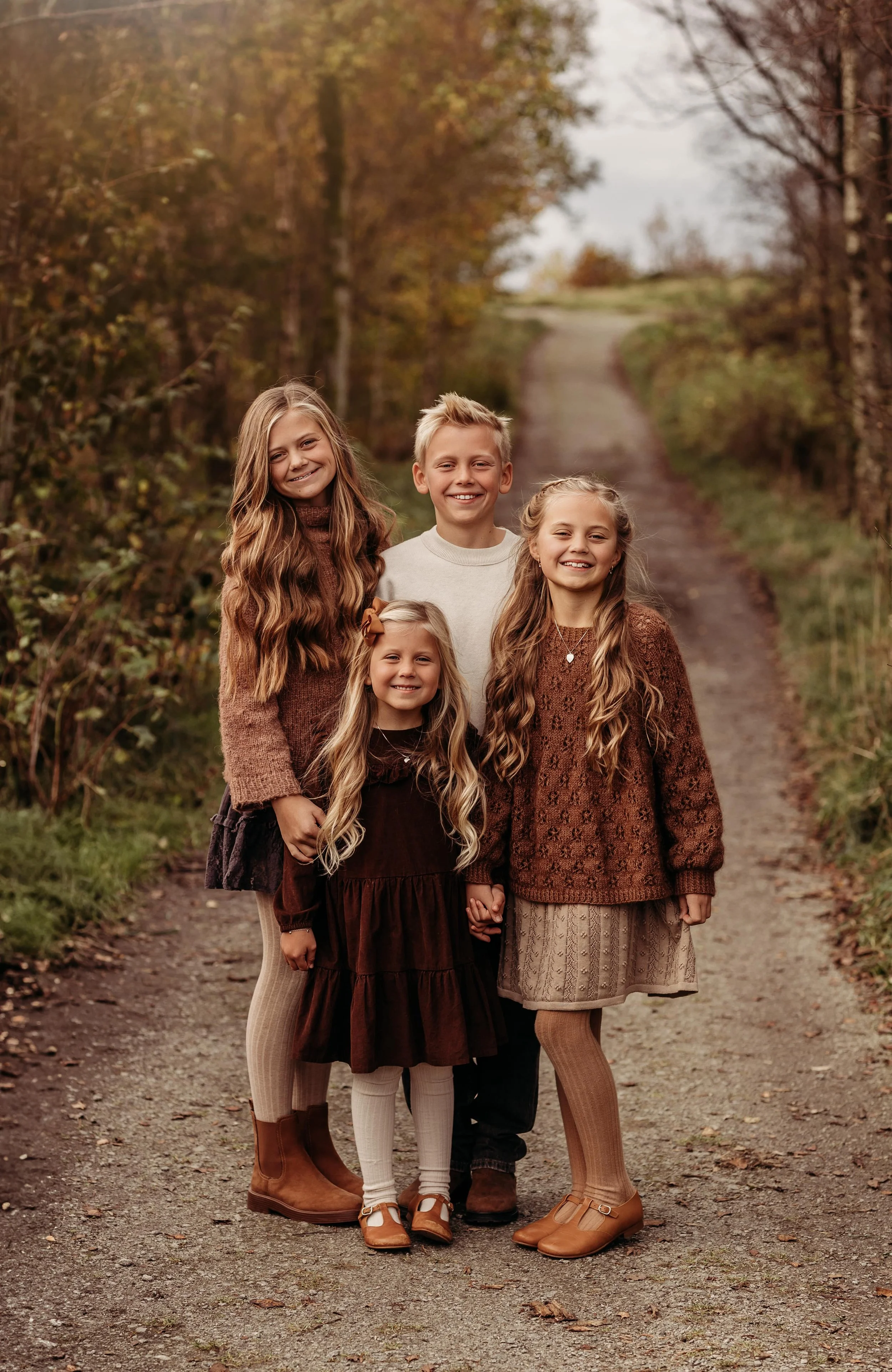 En gruppe på fem barn står på en grusvei i høstlandskap, omgitt av trær med høstfarger, smilende og holdende hender.