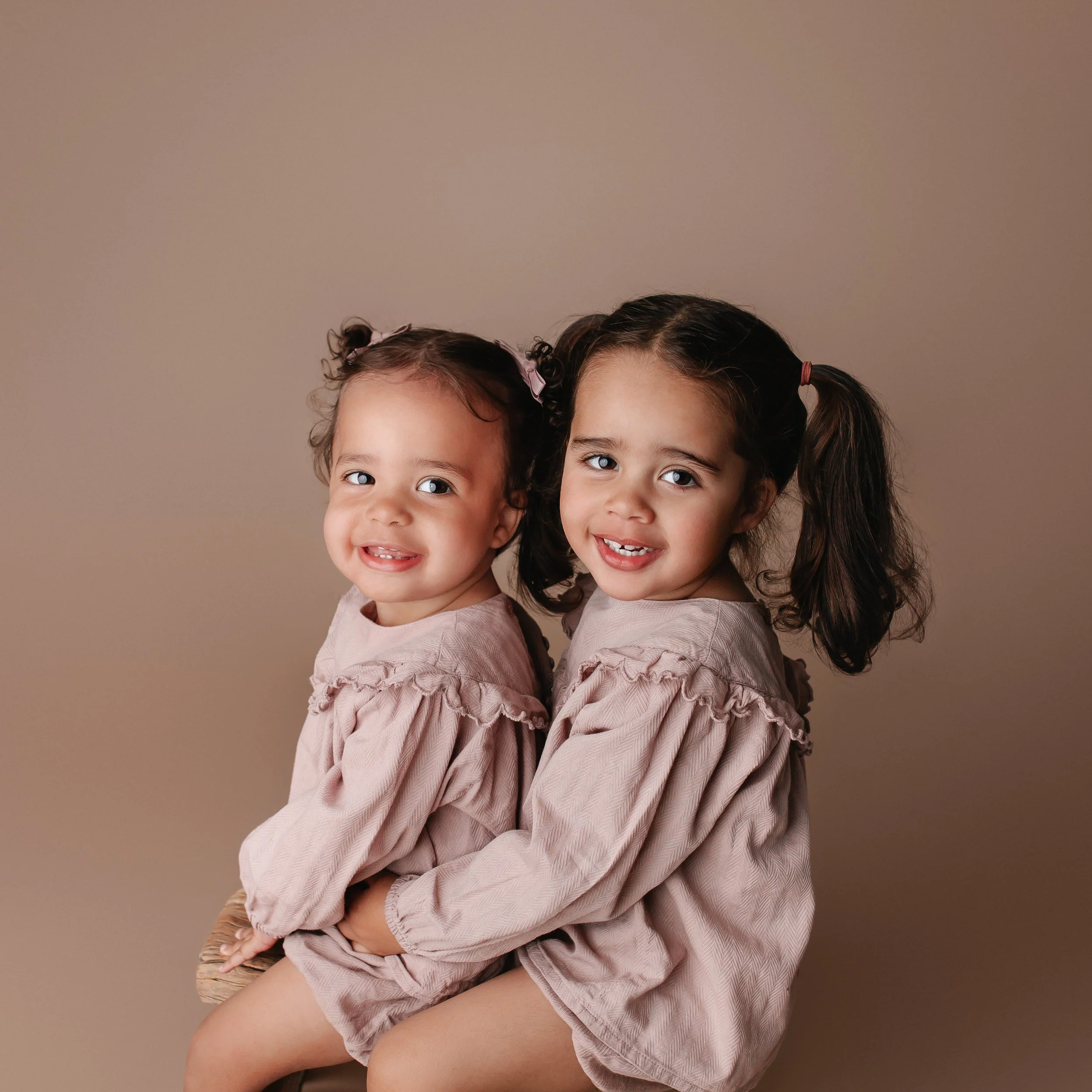 To unge jenter i rosa kjoler med pigtails, som smiler og sitter tett sammen mot en beige bakgrunn.