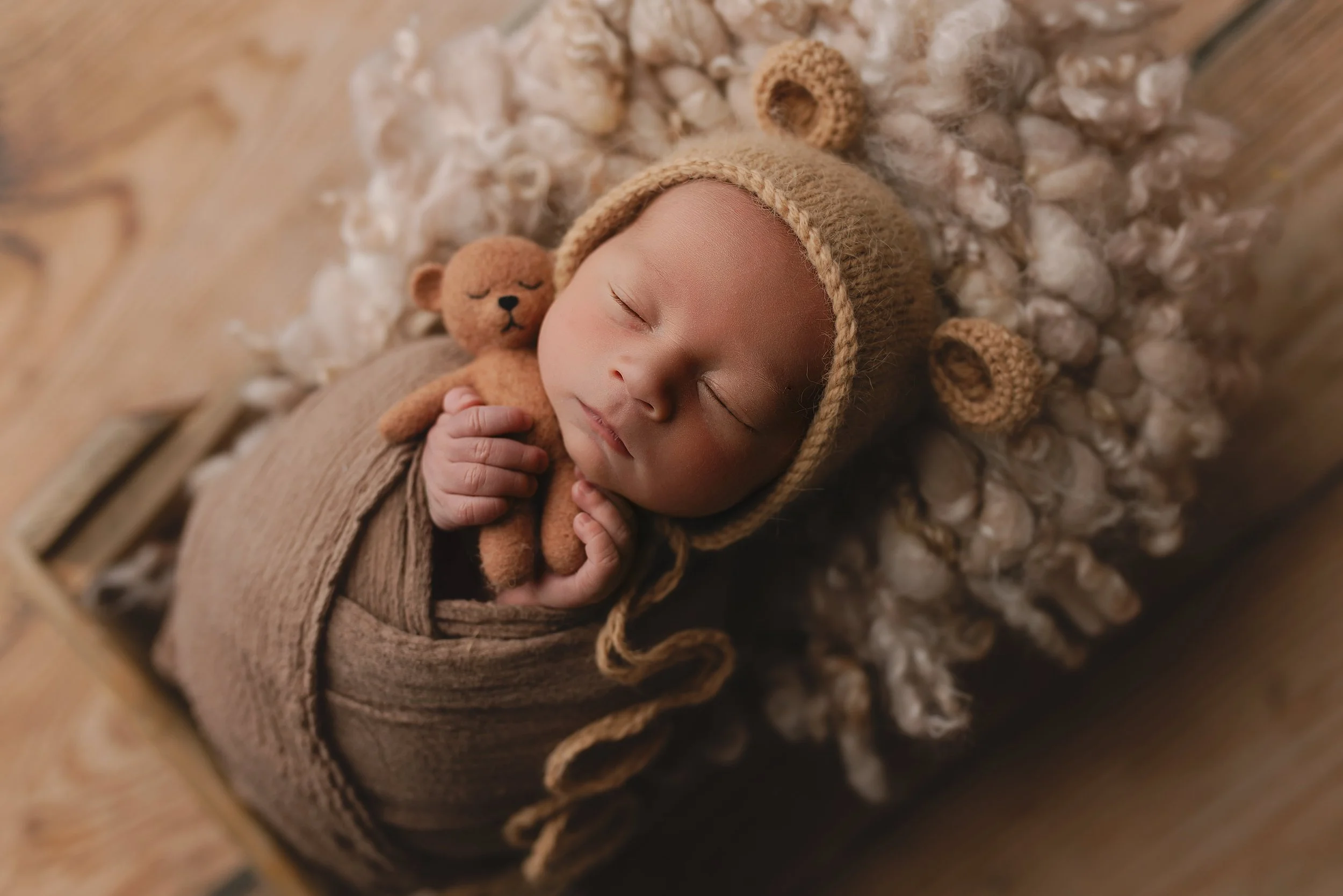 Søvnende baby med nisselue, gnedende en liten teddybjørn, liggende på en myk, kremfarget pels på et trebord