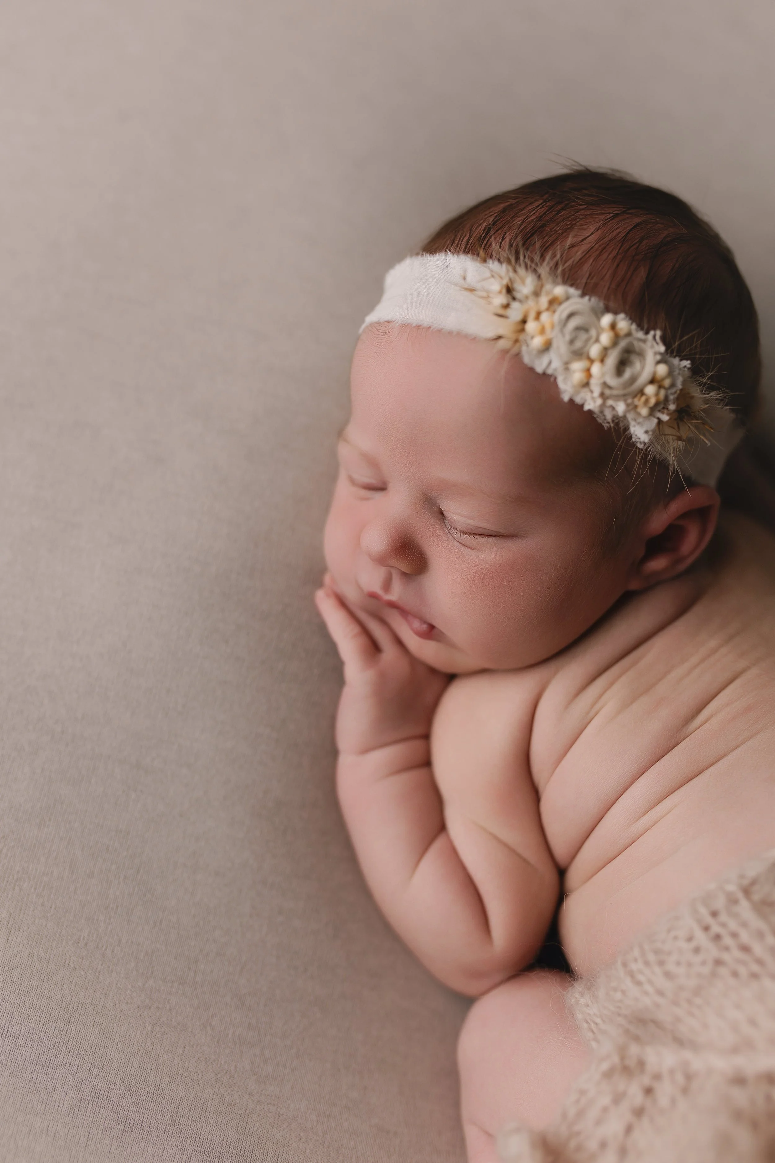 En nyfødt baby sover på en beige bakgrunn, har på seg en blomsterhodebånd.