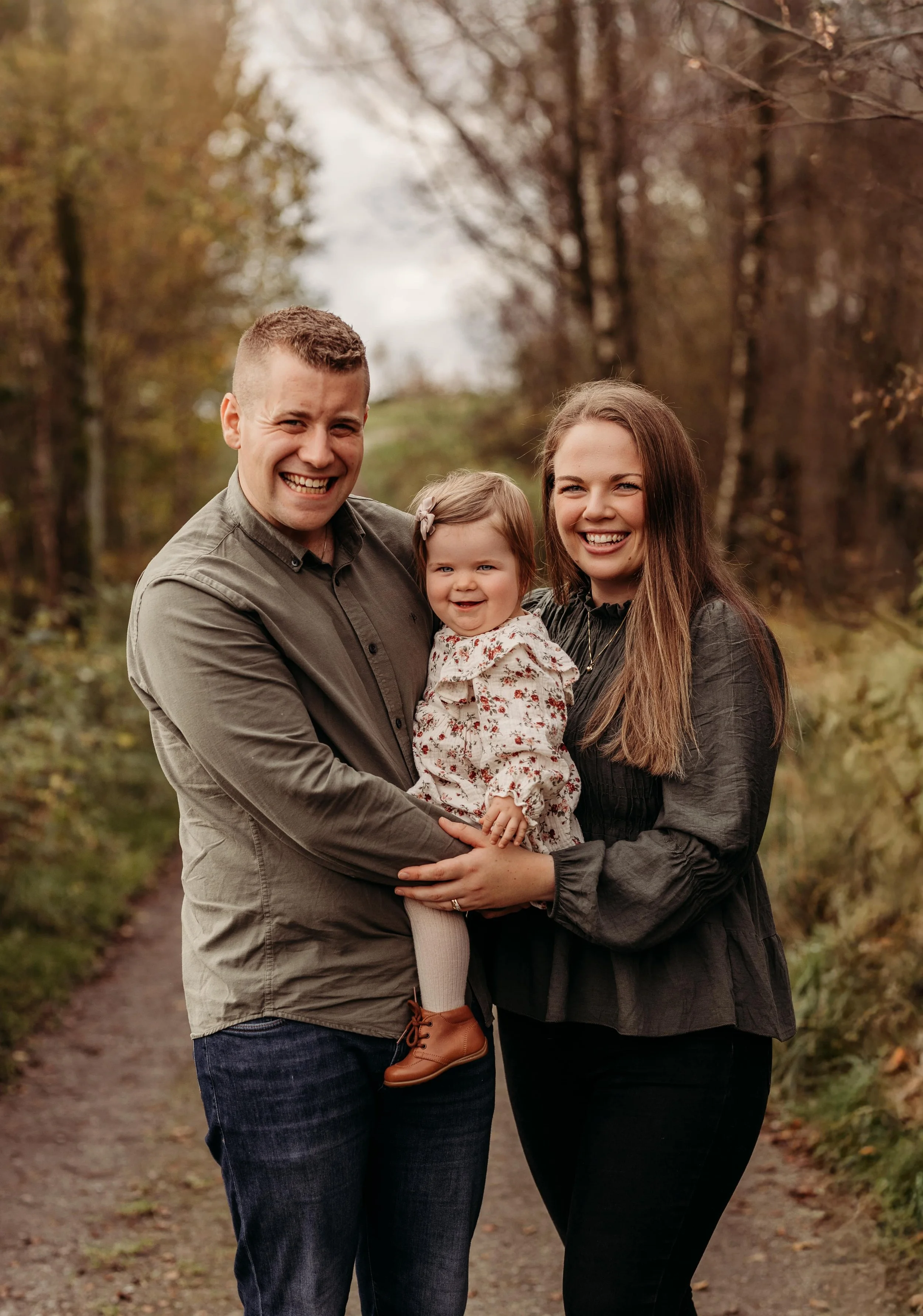 En lykkelig familie med to voksne og ett barn står på en skogssti under høst. De smiler og holder rundt hverandre.