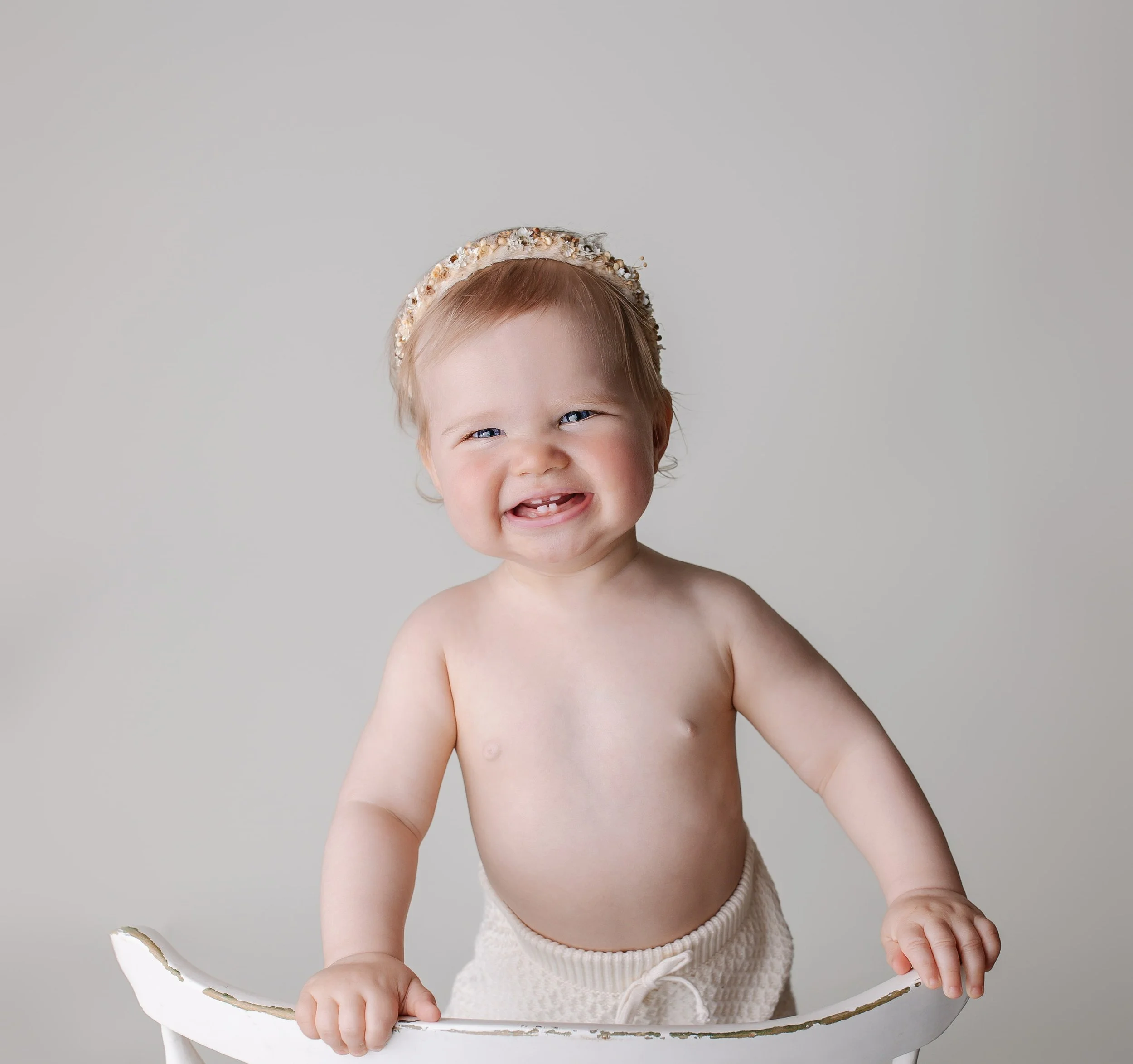 En smilende babyjente med blond hår og blå øyne, som holder på en hvit stol, med bare overkropp og beige bukser, mot en lys grå bakgrunn.