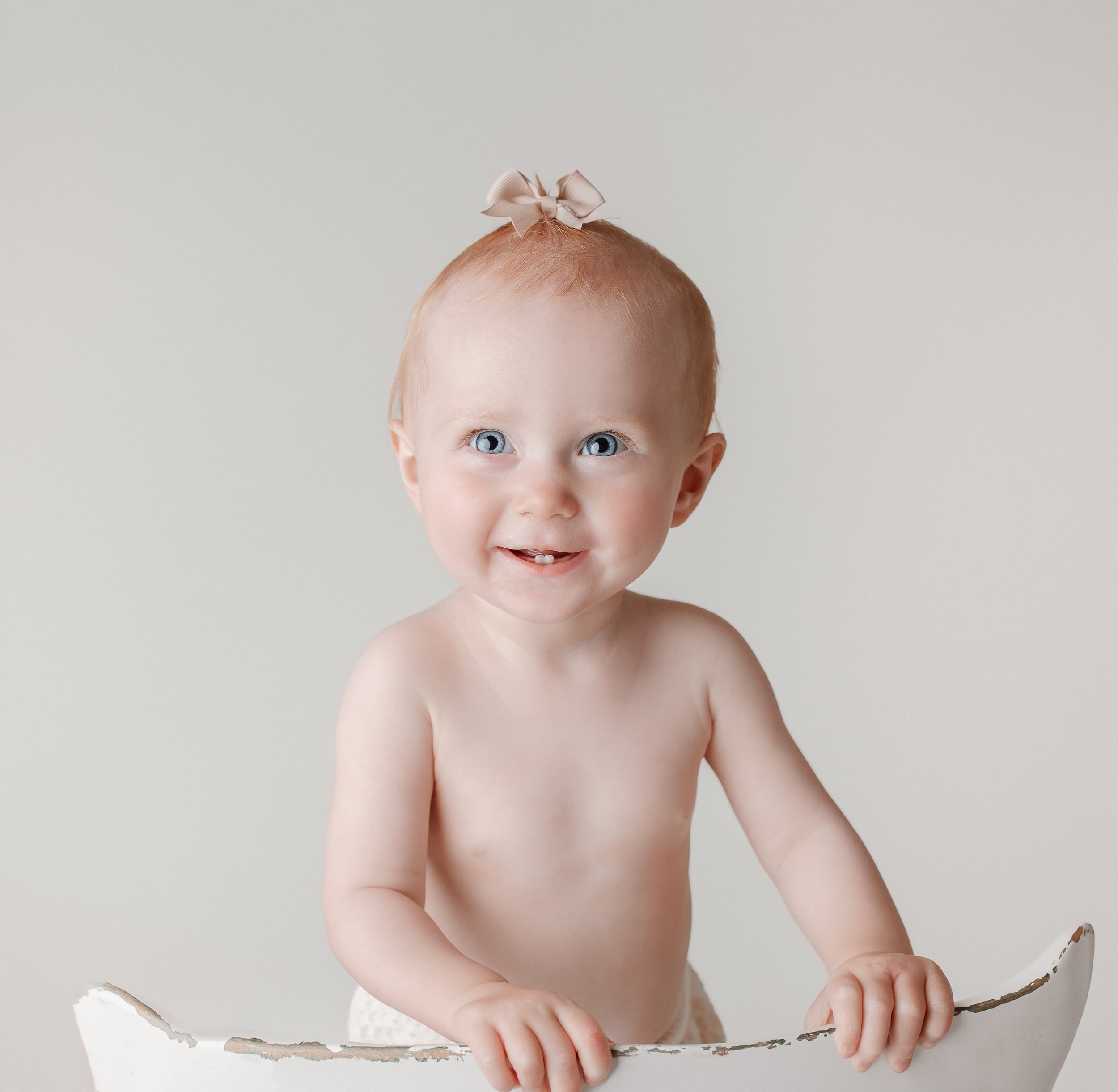 Søt, skrap barnehagegutt uten klær med blå øyne og rød hår, med en beige sløyfe i håret, som holder seg på en hvit leke, mot en enkel, lys bakgrunn.